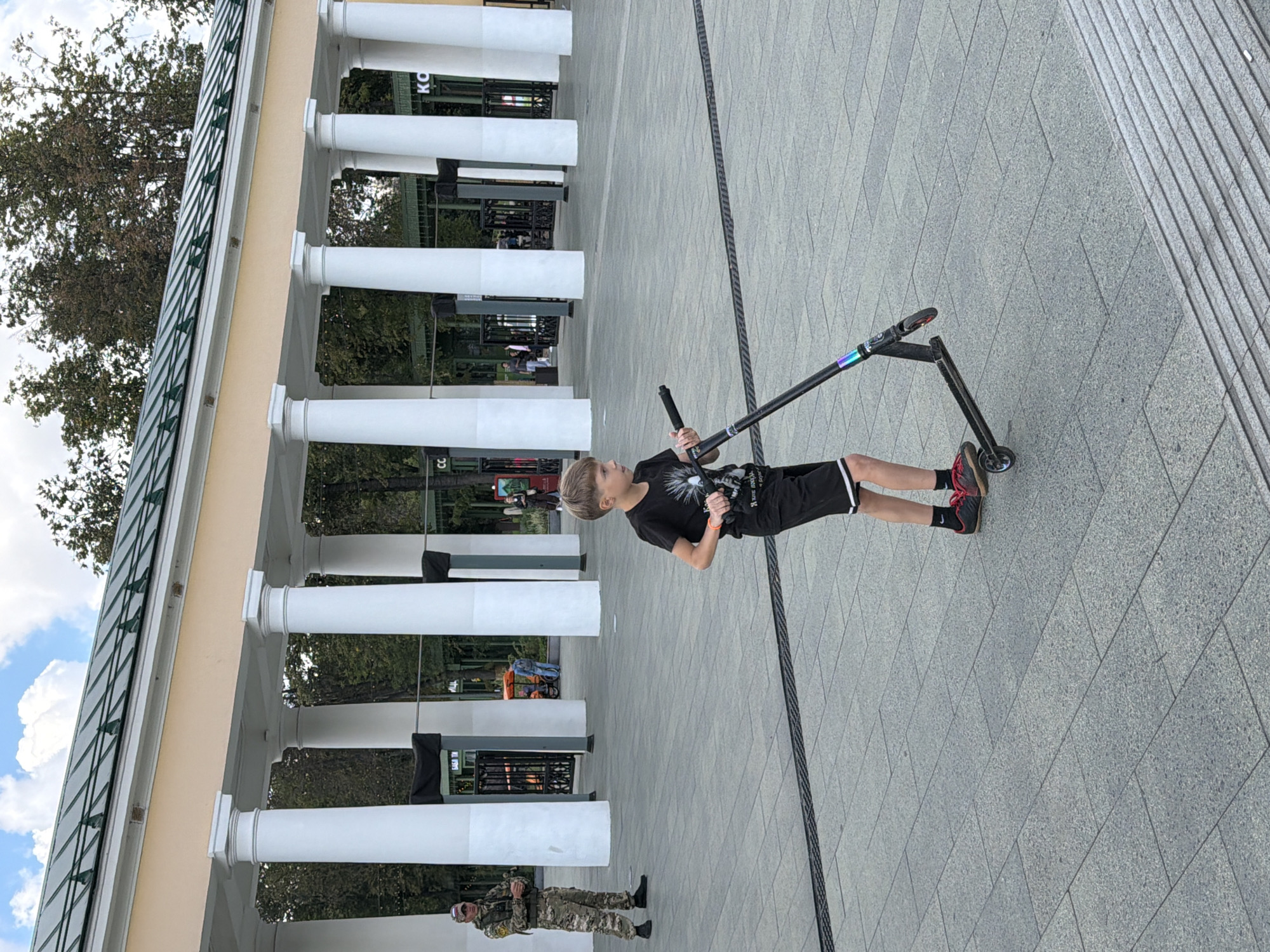 A young boy with blond hair, wearing a black t-shirt with a graphic print and black shorts with white stripes, rides a black scooter on a paved area. He is wearing red and black athletic shoes. To the left of the boy, a man in camouflage uniform stands at attention, facing the boy. Behind them, a colonnade of white pillars supports a light-colored awning with a green roof. The area appears to be an outdoor public space. The presence of the security guard and the architectural style suggest a park or a cultural institution in Nizhny Novgorod, Russia.