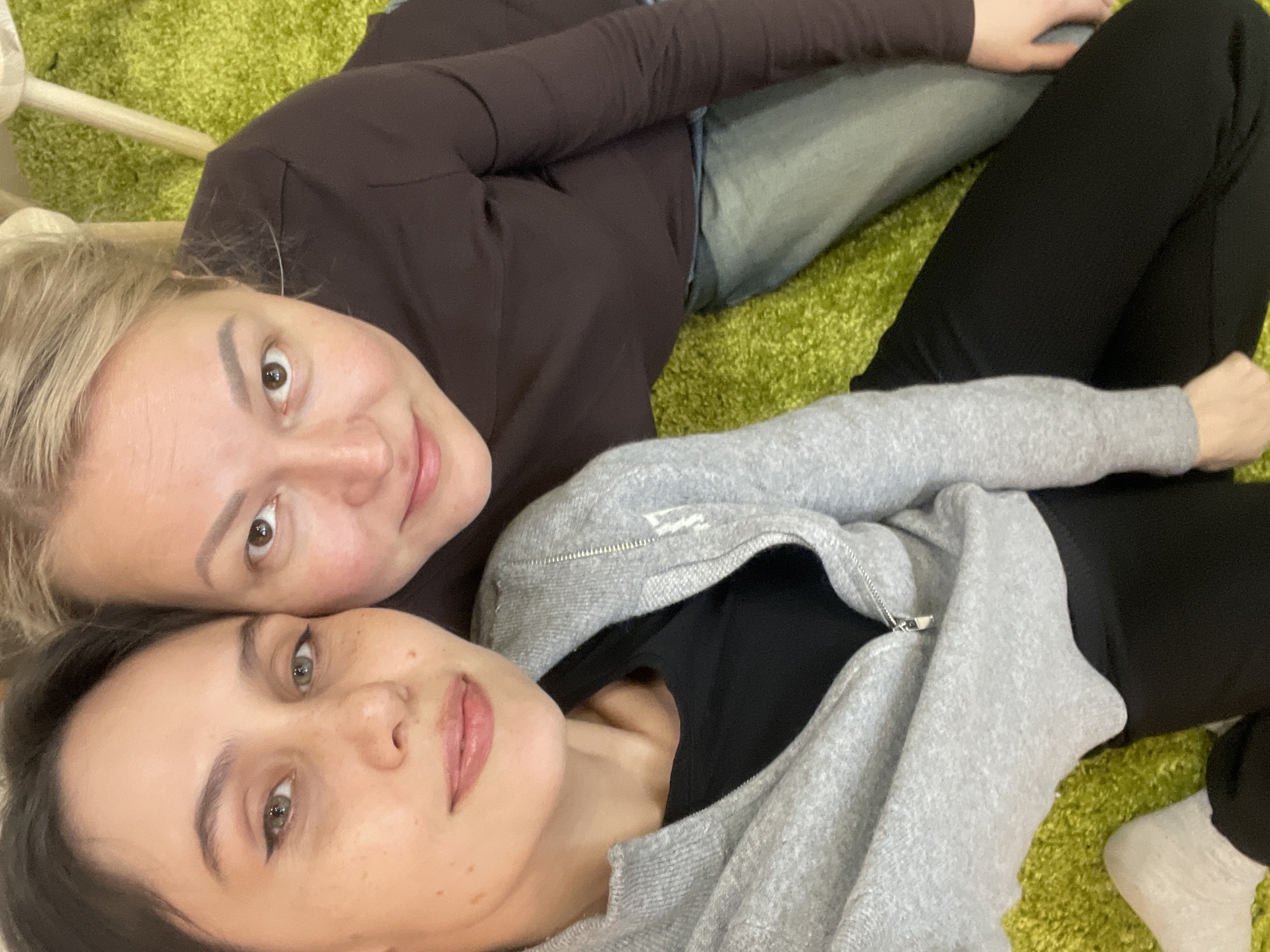 Two women are lying on a green rug indoors. The woman on top has blonde hair, a brown long-sleeved shirt, and blue jeans. The woman below has dark hair and wears a grey zip-up hoodie over a black shirt, along with black pants. The background is nondescript. The location is not identifiable as Izhevsk, Russia, from the information available in the image.