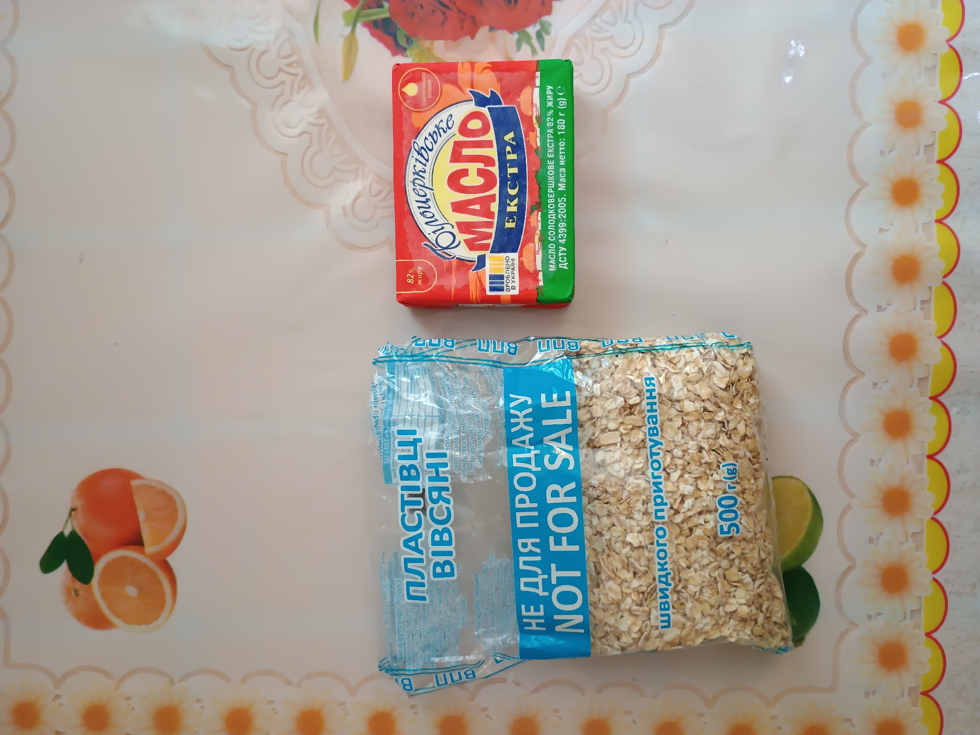 The image displays a tabletop scene featuring food items. The primary subjects are a block of butter and a bag of rolled oats, both common grocery items. The butter is packaged in a red wrapper with Ukrainian text and an illustration of orange slices. The rolled oats are in a clear plastic bag, also with Ukrainian text, and a prominent "NOT FOR SALE" label.
The background consists of a light-colored tablecloth with a decorative floral pattern along the right edge. In the lower left corner, there are several orange slices and a lime, suggesting a potential use or flavor association for the products. The lighting appears to be natural, possibly indicating daytime. There are no people present in the image, and the focus is solely on the arrangement of the food items.
The visible text on the butter wrapper includes "МАСЛО ЕКСТРА" (Butter Extra) and "82% жиру" (82% fat). The bag of oats has text like "ПЛАСТІВЦІ ВІВСЯНІ" (Rolled Oats), "НЕ ДЛЯ ПРОДАЖУ" (NOT FOR SALE), and "швидкого приготування" (quick cooking). The location context is provided as Zaporizhzhia, Ukraine. The overall scene is simple and domestic, suggesting a casual setting like a kitchen table.