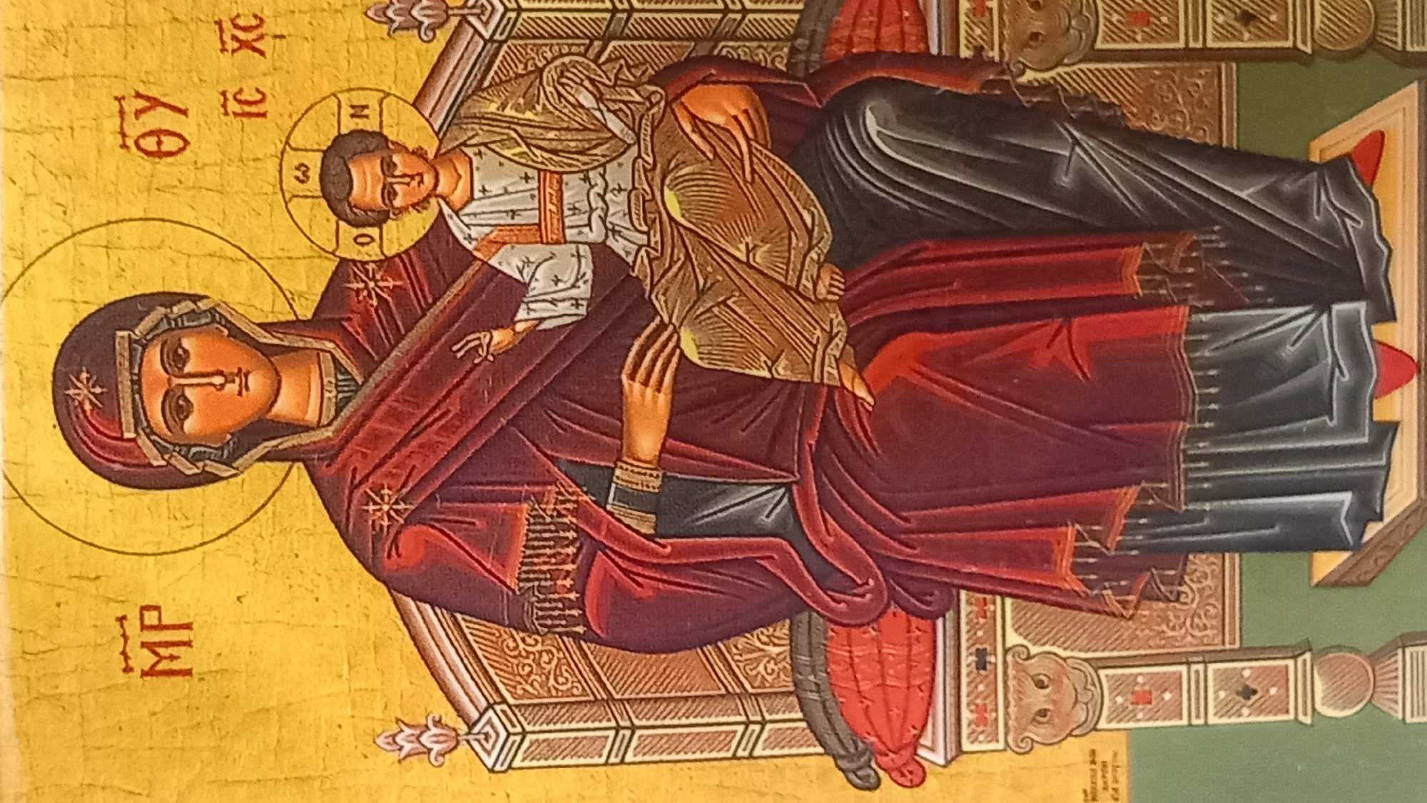 This is a close-up, full-frame photograph of an icon depicting the Virgin Mary holding the Christ child. The figures are rendered in a traditional Byzantine style with gold leaf background. The Virgin Mary is depicted seated on an ornate throne, wearing a dark red mantle over a dark blue garment. She has a halo with Greek letters and a starry decoration on her head covering. The Christ child is seated on her lap, also with a halo and Greek inscriptions. He is dressed in a white tunic and a golden mantle, and is making a blessing gesture with his right hand. The throne is richly decorated with intricate patterns. The overall impression is one of solemnity and reverence. The icon is located in the city of Kos, Greece.
