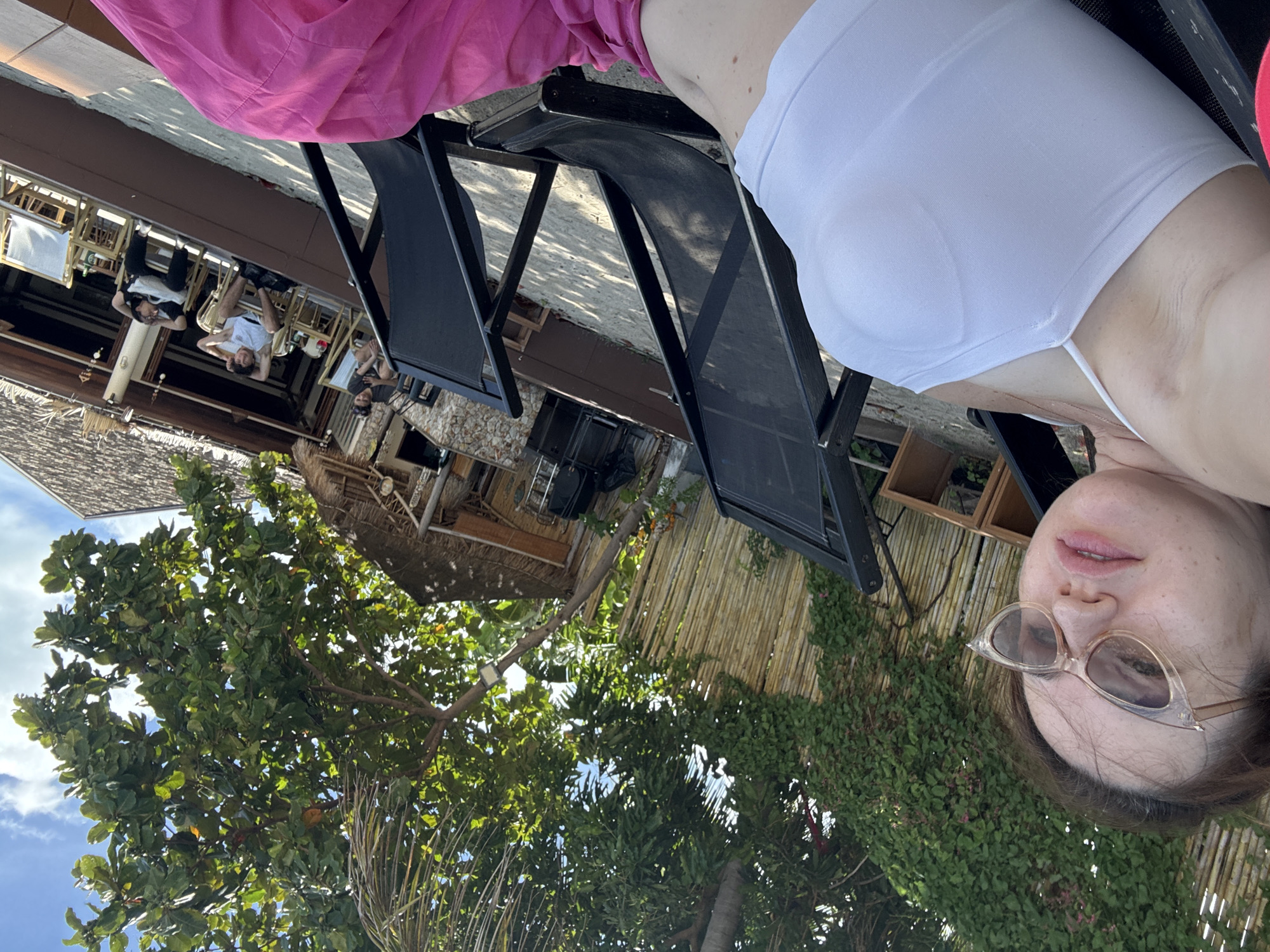 A woman is lying down, facing upwards, with her head towards the bottom right of the frame. She is wearing a white top and large, cat-eye sunglasses. Her lips are slightly parted. Behind her, to the left, a pink garment is visible. The scene appears to be outdoors, with lush green foliage and trees in the background, suggesting a tropical setting. A wooden structure with a thatched roof is also visible, along with some black lounge chairs. The lighting suggests it is daytime, possibly late afternoon given the shadows. The overall mood seems relaxed and summery. The image is oriented sideways.