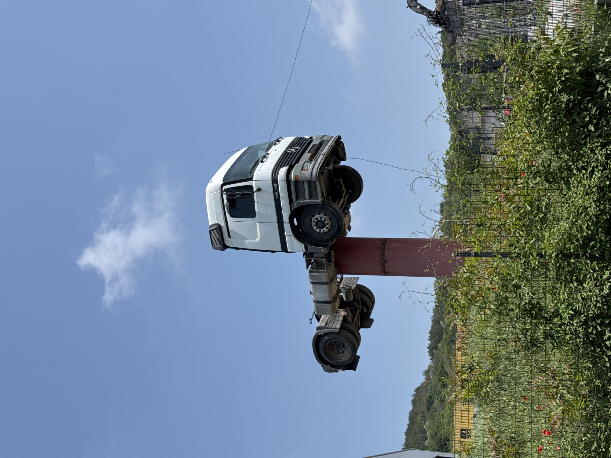 A white semi-truck cabin and chassis are prominently displayed, mounted vertically on top of a tall, red cylindrical column. The truck's cab rests on the column, with its front grille facing upwards and the chassis extending downwards towards the ground. The side of the truck's cab clearly shows the number "1835," and the Mercedes-Benz logo is visible on the front grille. Thin wires are noticeable near the front of the truck.
The scene is set outdoors under a bright blue sky with a few sparse white clouds, indicating daytime. In the background, to the right and bottom of the truck, there is a dense expanse of green foliage and bushes, partially obscuring a chain-link fence. Beyond the fence, more greenery covers what appear to be distant hills or a forested area. The overall setting suggests a rural or semi-rural location.
The arrangement of the truck on the pole appears to be a static display, possibly an art installation, a landmark, or an advertising structure. There is no discernible activity taking place, and no people are visible in the image. The weather is clear and sunny.
