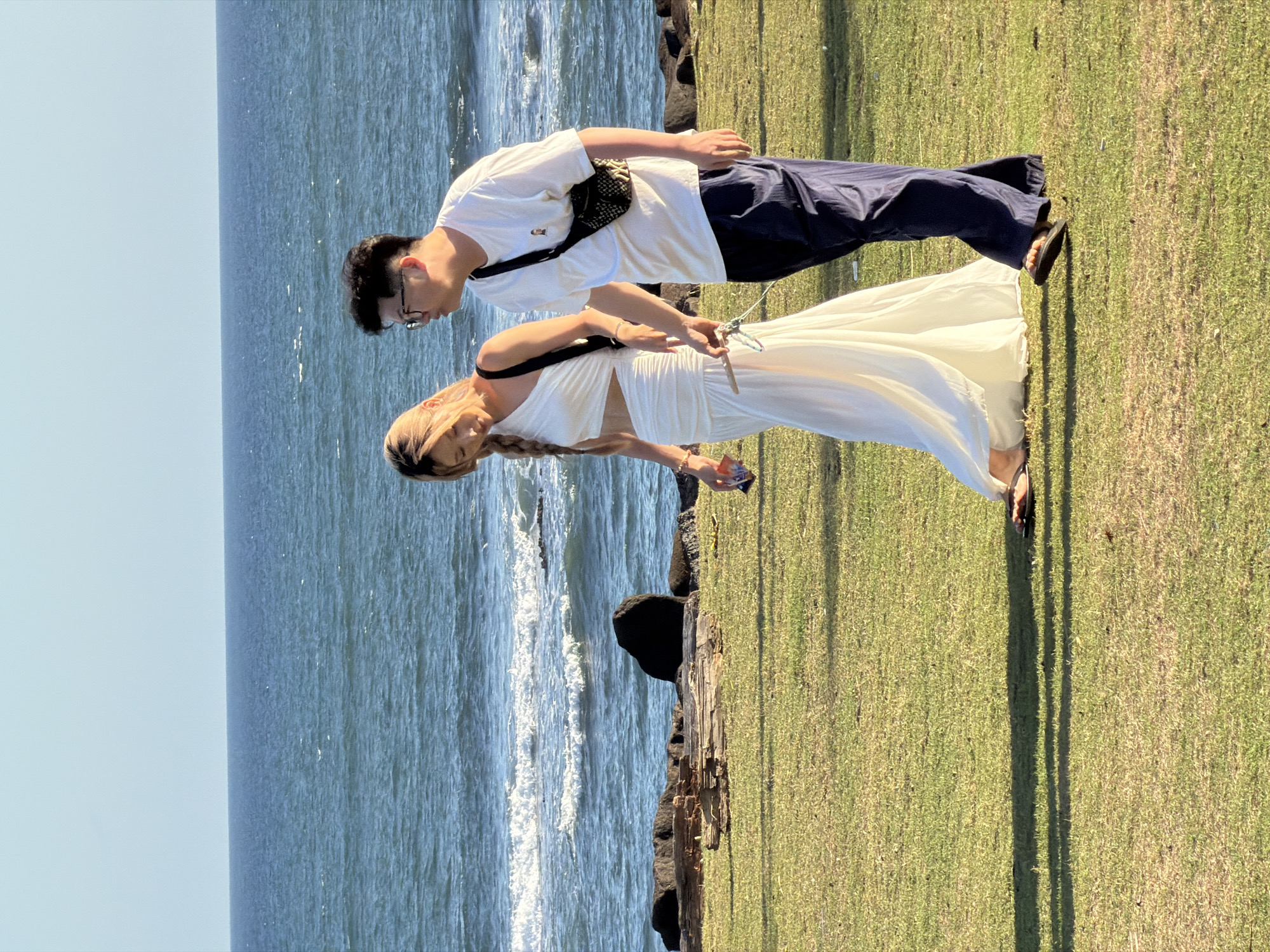 A young Asian couple is walking on a grassy area near the ocean. The woman, on the left, is wearing a white, cut-out, sleeveless dress and has her hair in a braid. The man, on the right, is wearing a white t-shirt, dark pants, and glasses. He has a dark crossbody bag over his shoulder and a patterned fanny pack around his waist. They are walking towards the right side of the frame and appear to be holding hands. The ocean is visible behind them, with waves breaking on the shore. There are also some rocks and driftwood on the shoreline. The location is the city of Mengwi, Indonesia.