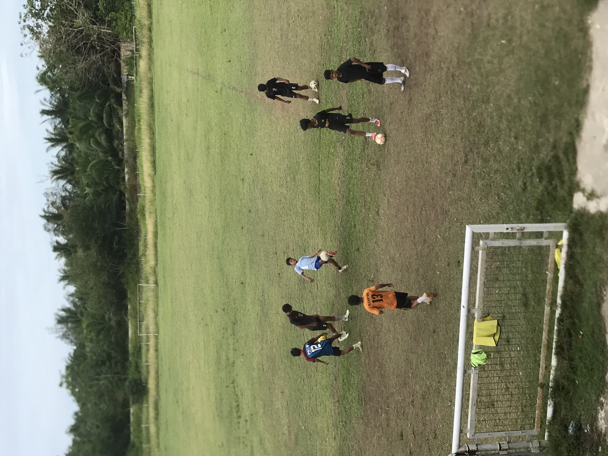 The image shows a soccer match taking place on a grass field. Seven people are visible in the field. Some of the players are wearing black jerseys, one is wearing orange, and one is wearing light blue. One goal is visible, with two yellow soccer vests and a neon green soccer ball near the goal. The photo was taken in Palohlada, Indonesia, as evidenced by the text on the player's orange jersey. The vegetation on the left-hand side indicates a tropical location.