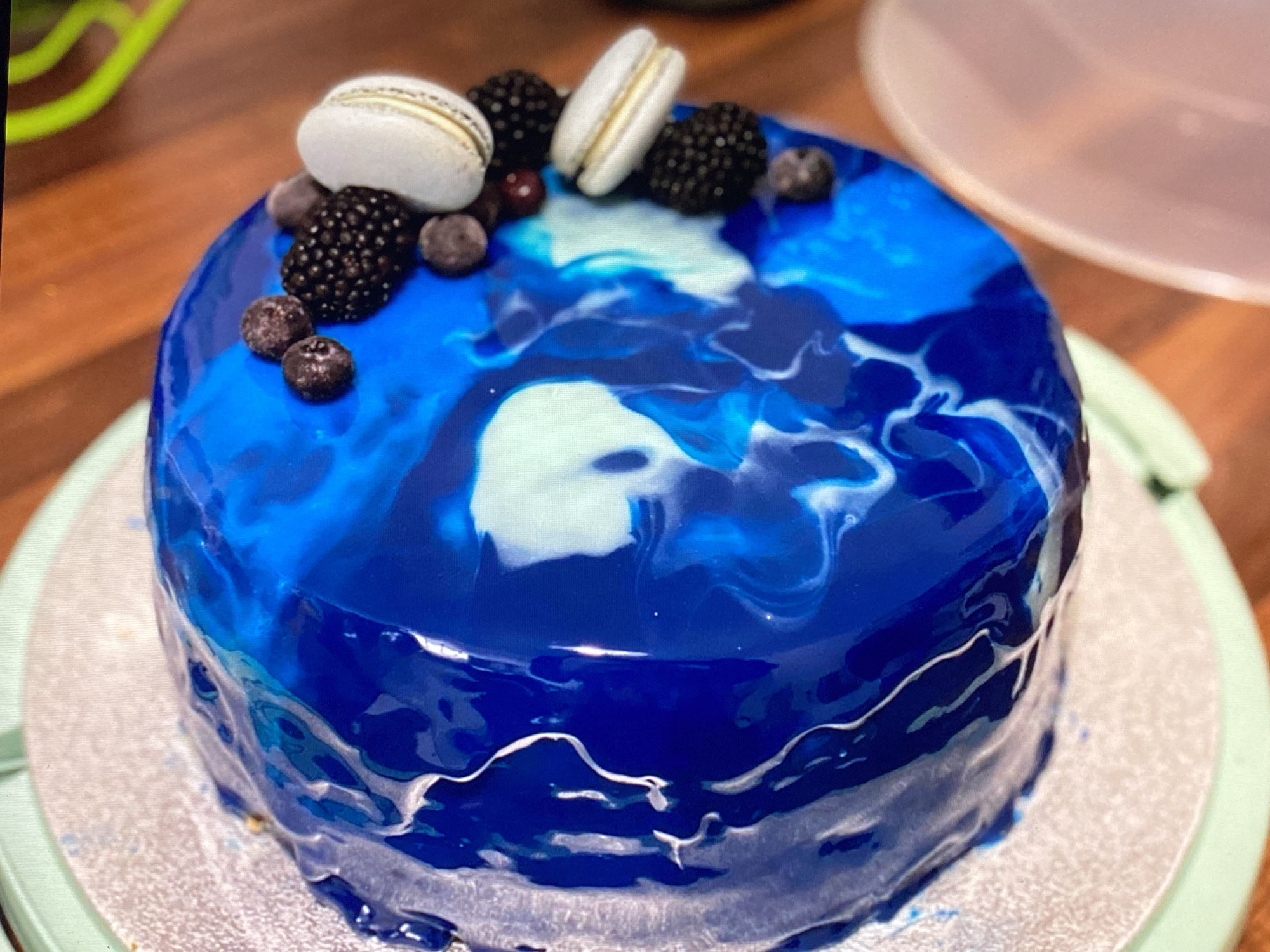 The image shows a close-up of a round cake with a glossy blue and white marbled glaze. The cake is decorated with several fresh blackberries, blueberries, and two white macarons. It is sitting on a silver cake board, which is on a light green cake stand. The background is a blurred wooden surface and a glimpse of a light pink bowl. There is no indication of any specific location in Purkersdorf, Austria, or any people in the image.