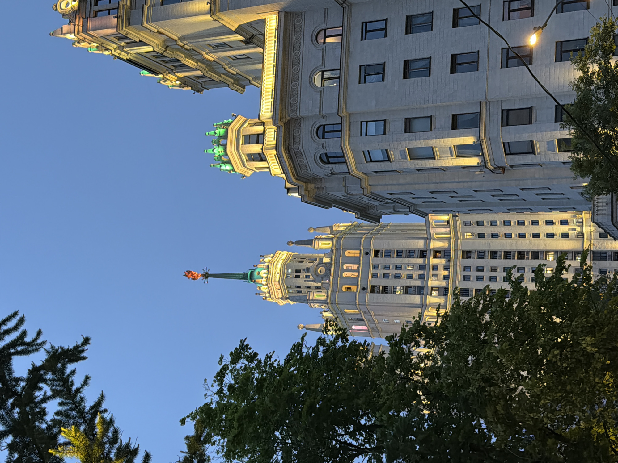 The image shows a low-angle view of two illuminated, monumental buildings against a clear blue sky at dusk or dawn. The building in the upper left corner features elaborate architectural details and green decorative elements. The second building, positioned towards the bottom center of the frame, is taller and topped with a green spire and a red star-like structure. Tree foliage is visible in the foreground, partially obscuring the lower portions of the second building. A street light and its cable are visible on the right side of the image.
The location is not in Перовское сельское поселение, Ukraine, since the buildings are examples of Stalinist architecture in Moscow, Russia. They are some of the Seven Sisters.