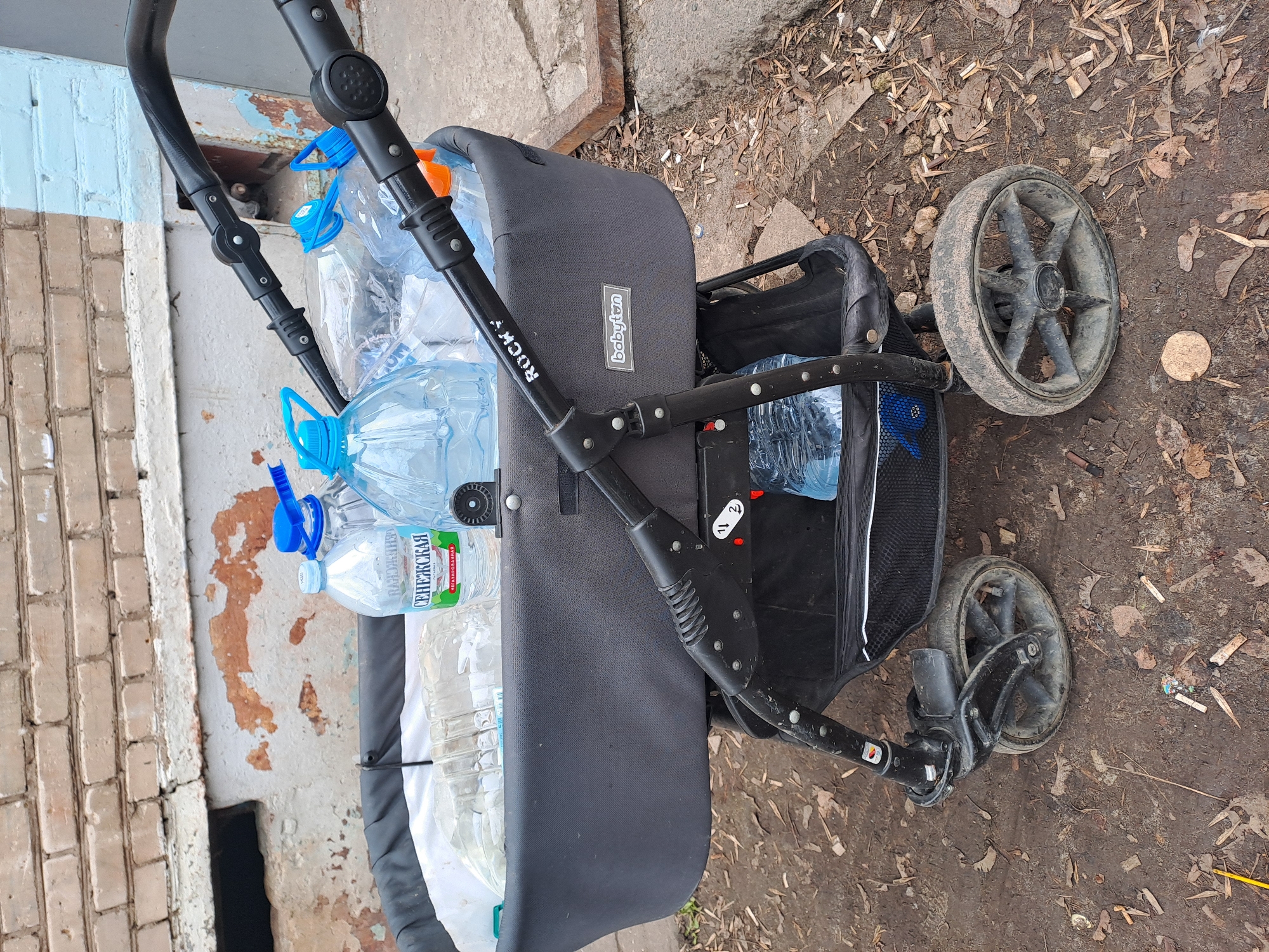 The image shows a baby stroller parked near a brick building in what appears to be an urban outdoor environment. The stroller is grey and black, filled with several clear plastic bottles of water. The brand name "babyton ROCKY" is visible on the side. The wheels of the stroller are dusty. The ground around the stroller is dirt with scattered leaves. The building next to the stroller is weathered with peeling paint and brickwork. The side of the building is next to a brick wall.
The location is Elektrostal, Russia, based on the inscription on the bottle.