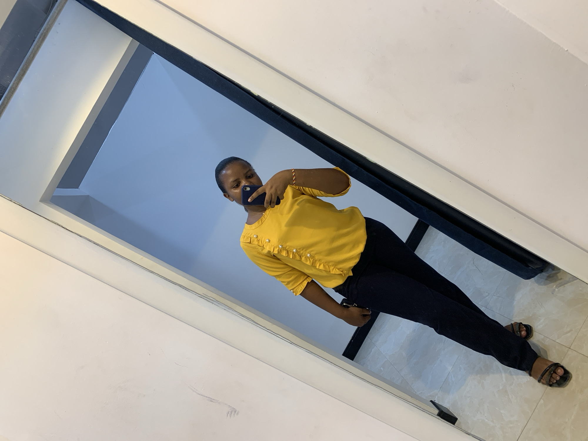 A person stands in front of a mirror taking a selfie. The individual wears a yellow top with ruffles and pearl embellishments, dark-colored pants, and sandals. They hold a mobile phone in their hand, partially obscuring their face. The mirror reflects an interior space, possibly a room with light blue walls and a white ceiling. The background appears to be a simple indoor setting. Without additional context or visual cues, it is impossible to confirm the location of this photograph in Dar es-Salaam, Tanzania.
