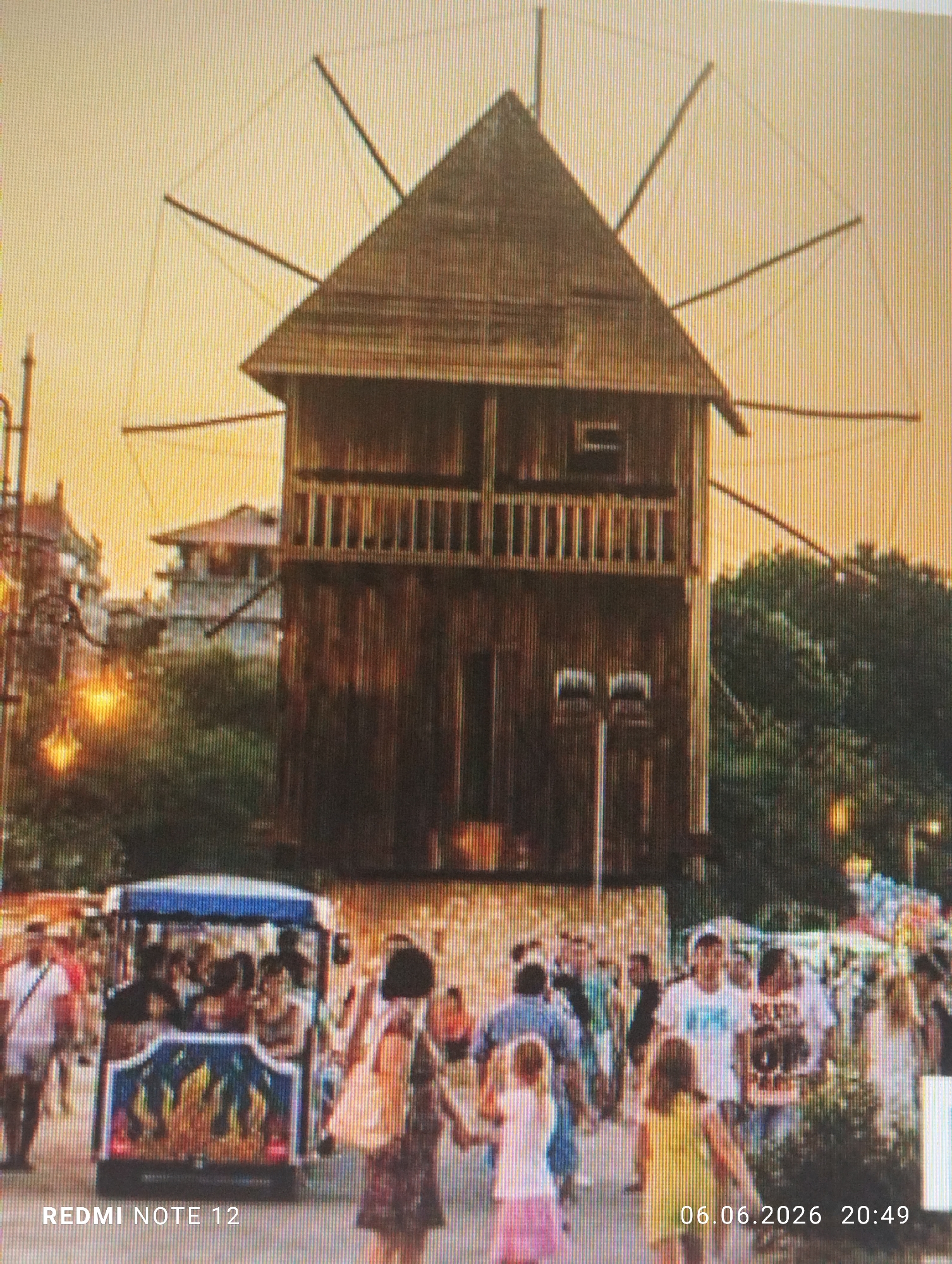 The image depicts the historic Old Windmill in Nessebar, Bulgaria, a town located approximately 35 kilometers north of Burgas, during twilight or early evening. The prominent central subject is a multi-story wooden windmill with a gabled roof, featuring a second-story balcony with balusters and multiple radiating wooden spokes, appearing as a static landmark rather than an operational mill. The structure exhibits a weathered, dark brown finish.
The foreground is a bustling pedestrian area populated by numerous individuals of varying ages, including adults and children, walking in multiple directions. On the left side, an open-sided tourist train or shuttle, painted with blue and red flame-like designs, is visible and occupied by several passengers. Artificial streetlights emit a warm, golden glow, indicating the onset of night. The sky transitions from orange to yellow hues, characteristic of sunset. In the midground and background, dense green foliage and outlines of other buildings are discernible.
The image contains a "REDMI NOTE 12" watermark in the bottom-left corner and a timestamp "06.06.2026 20:49" in the bottom-right corner. The overall image quality shows pixelation and vertical banding, suggesting it may be a photograph of a digital display.