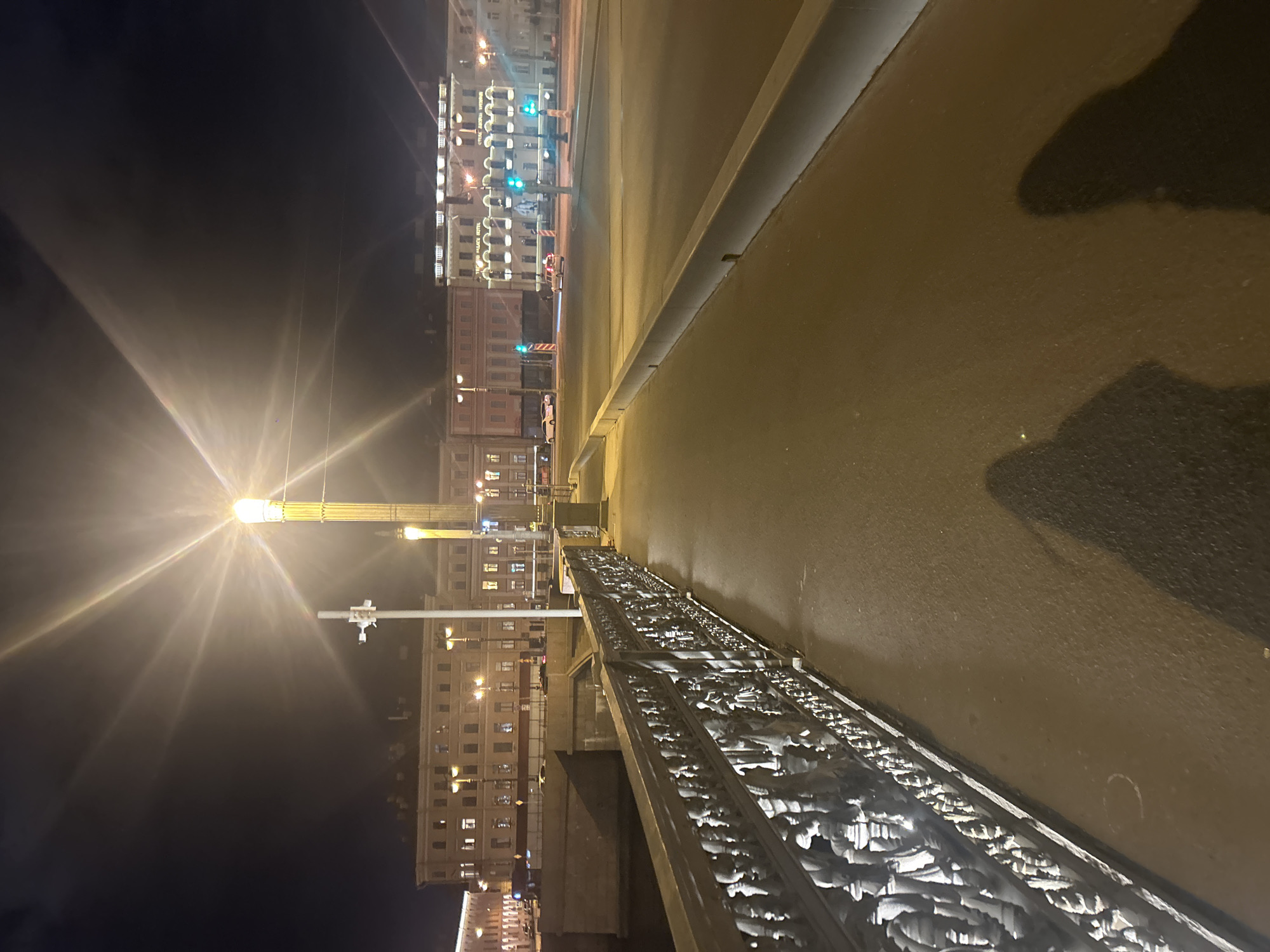 This media file captures a night scene in Saint Petersburg, Russia, from an elevated perspective, possibly a bridge. The image is rotated 90 degrees clockwise, placing the street and buildings on the right side of the frame and the sky on the left.
In the foreground, a dark, ornate bridge railing with intricate decorative patterns runs diagonally. Next to the railing, the road surface appears wet and reflective, suggesting recent rain or dampness, and shows dark, amorphous shapes, likely shadows or reflections.
The midground and background feature a city street lined with multi-story buildings. Many windows in these buildings are illuminated, suggesting activity or decorative lighting. Visible text on one building clearly reads "DOMINA ST.PETERSBURG" and "HOTEL," while another prominent building displays "ASTORIA HOTEL," strongly confirming the Saint Petersburg location.
Bright streetlights cast prominent starburst flares across the dark night sky, with one tall lamppost on the left side of the frame being particularly luminous. Traffic lights are visible further down the street, showing green and red signals. A white van with roof lights and another white car are also present on the street. The scene depicts a quiet, illuminated urban environment at night, with no direct human activity visible, but the lights and vehicles suggest ongoing city life.