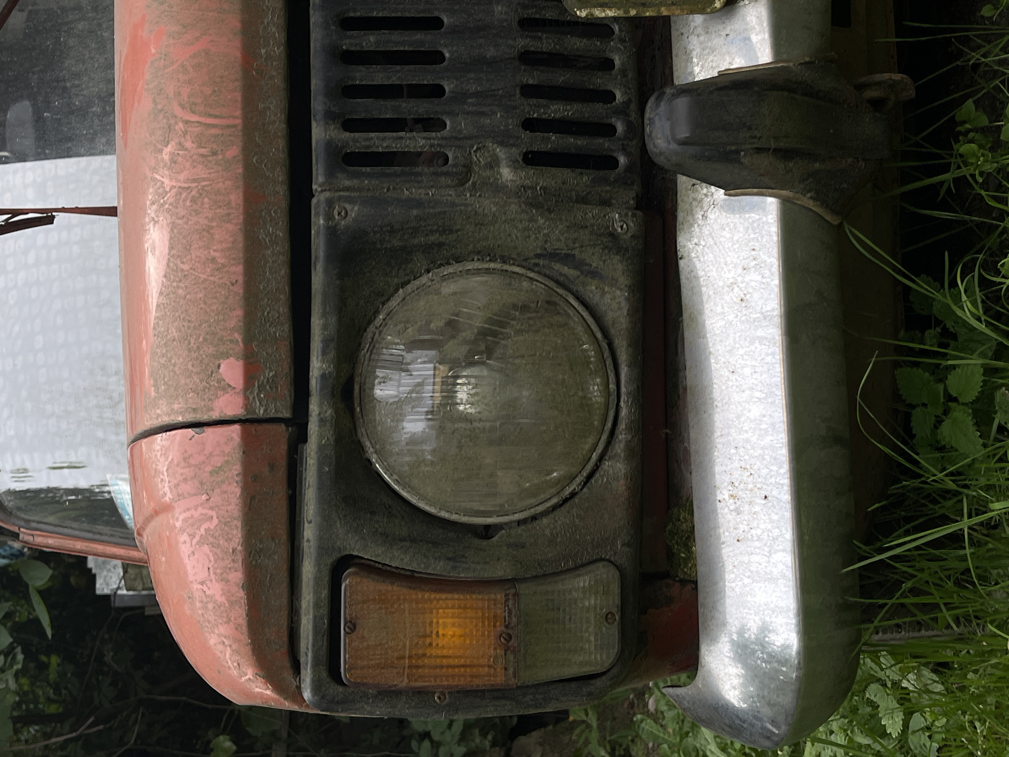The image shows a close-up view of the front of a weathered, red tractor. The tractor is partially covered in dust and dirt, indicating it has likely been used outdoors for some time.
The subjects in the image are primarily mechanical components of the tractor. A prominent circular headlight, with a cloudy lens, is centered in the frame. Below the headlight, there is a combined turn signal and parking light assembly, with an amber lens on the left and a clear lens on the right. Above the headlight, a black grille with horizontal slots is visible.
To the left of the headlight and grille, a portion of the tractor's red fender is visible. The paint appears faded and streaked with dust. To the right of the headlight, a shiny chrome bumper curves around the front of the tractor. The bumper is also slightly dusty.
The setting appears to be outdoors, with green foliage and grass visible in the background, suggesting a rural or overgrown environment. The lighting is somewhat diffused, but there are no strong shadows, which could indicate an overcast day or a time of day when the sun is not directly overhead.
There are no people or animals visible in the image, and no specific activity or event is taking place. The image focuses solely on the details of the tractor's front end. The overall impression is one of age and disuse, with the tractor showing signs of wear and tear from exposure to the elements. The location context of Lviv, Ukraine, does not provide further visual cues within the image itself. No visible text is present.
