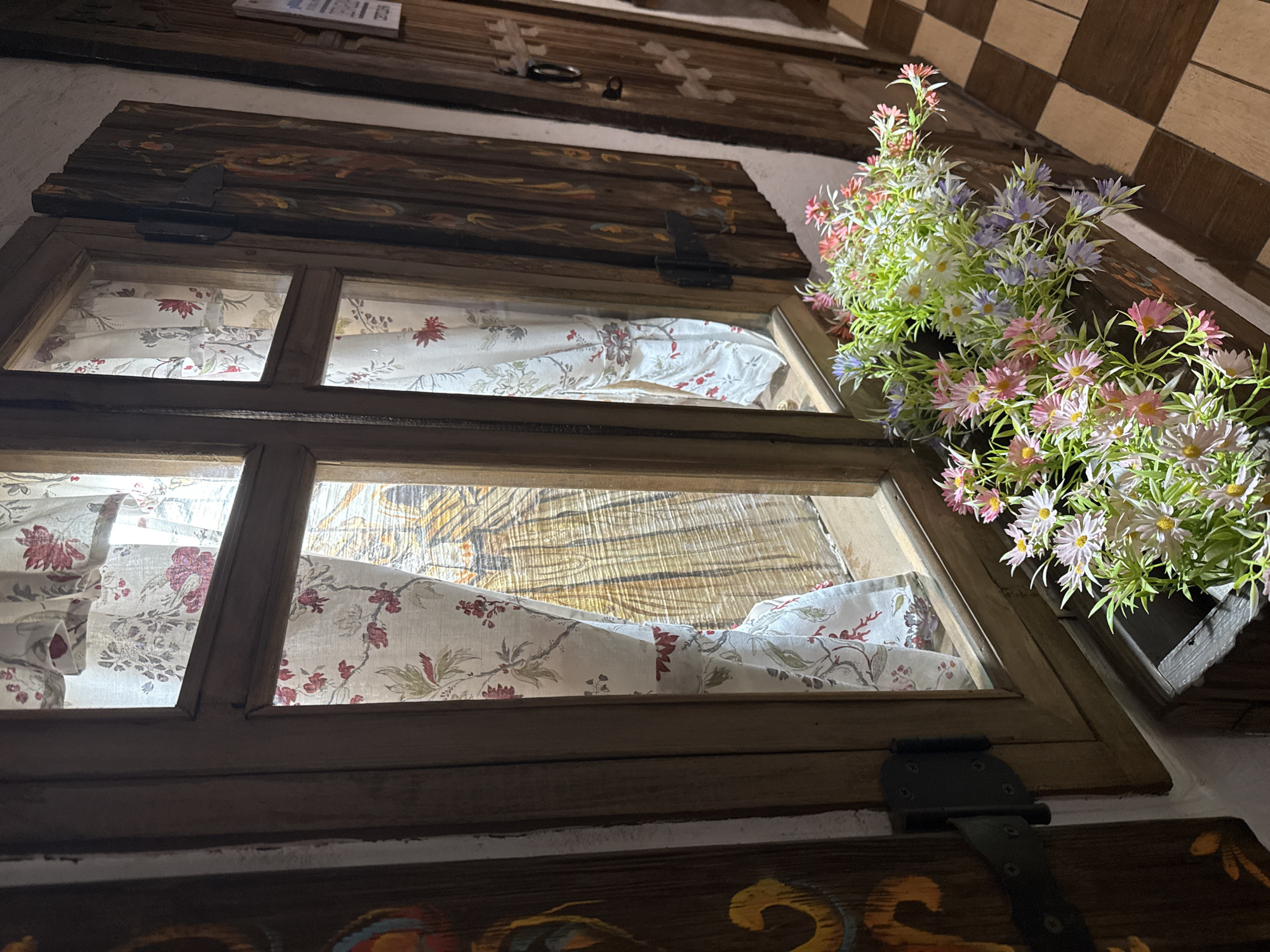 This is a medium shot, eye-level, outdoor photograph of a rustic wooden window with decorative shutters and a flower box. The window is divided into two panes by a wooden mullion. The shutters are ornately carved and painted with colorful, abstract designs. They are partially open, revealing floral patterned curtains behind the glass. A wooden planter box filled with an assortment of artificial flowers in shades of pink, white, and purple is placed on the windowsill below the window. The flowers are brightly lit, contrasting with the darker wood and stone elements of the scene. In the background, a checkered floor made of brown and beige tiles is visible. A wooden door with a metal handle and decorative carvings is situated to the right of the window. The overall scene suggests a charming and quaint architectural detail, possibly in a village or older part of a city, given the rustic aesthetic. The location is specified as Noviy, Russia. There are no people or animals present in the image, and no discernible actions or interactions are taking place. The lighting is uneven, with some areas brightly illuminated by an unseen light source, and others in shadow, creating a dramatic effect.