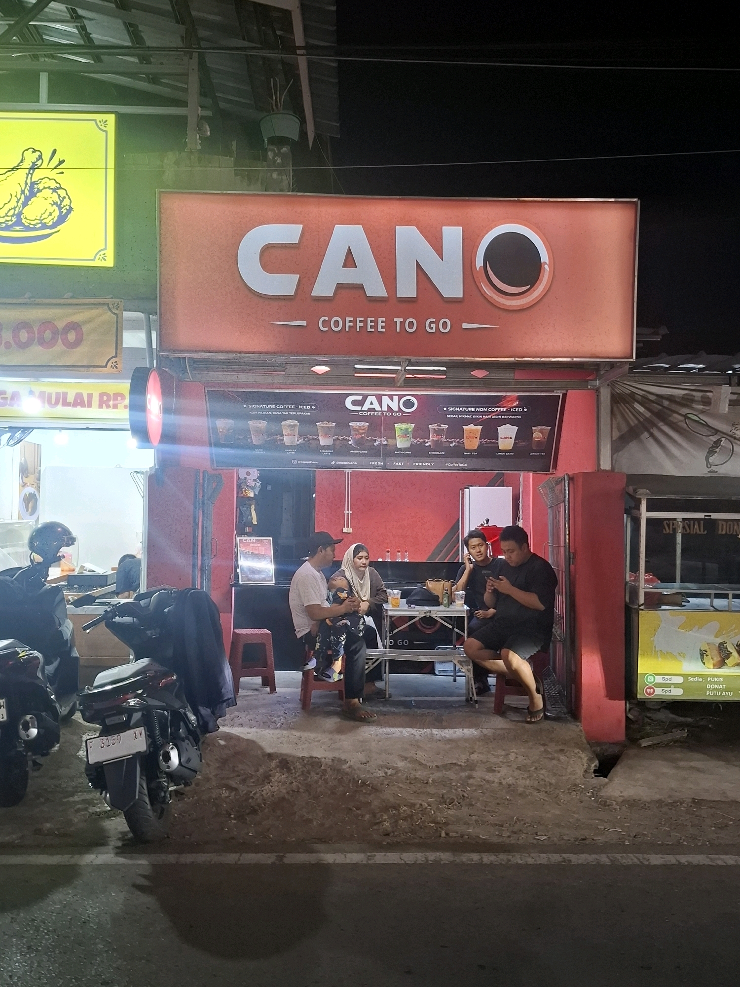 The image depicts a street-level view of a "CANO COFFEE TO GO" shop in Muka, Indonesia. The shop has a prominent orange sign with white lettering that reads "CANO COFFEE TO GO". Below the main sign, a smaller sign displays images of various coffee drinks and the text "SIGNATURE COFFEE - ICED" and "SIGNATURE NON COFFEE - ICED".
In front of the shop, several people are gathered. Two men and a woman are seated at a small table with drinks. A woman is holding a baby, and another man is seated to the right, looking at his phone. A child is also present, sitting on the lap of the woman holding the baby. Two motorcycles are parked on the left side of the image, one of which has a license plate with the number "F 3159 XY".
The environment appears to be an outdoor setting at night, with artificial lighting illuminating the scene. There are other shops and signs visible in the background, including a bright yellow sign with a drawing of fried chicken on the left and a sign for "SPESIAL DONUT" and "PUTU AYU" on the right. The ground is a mixture of dirt and asphalt.