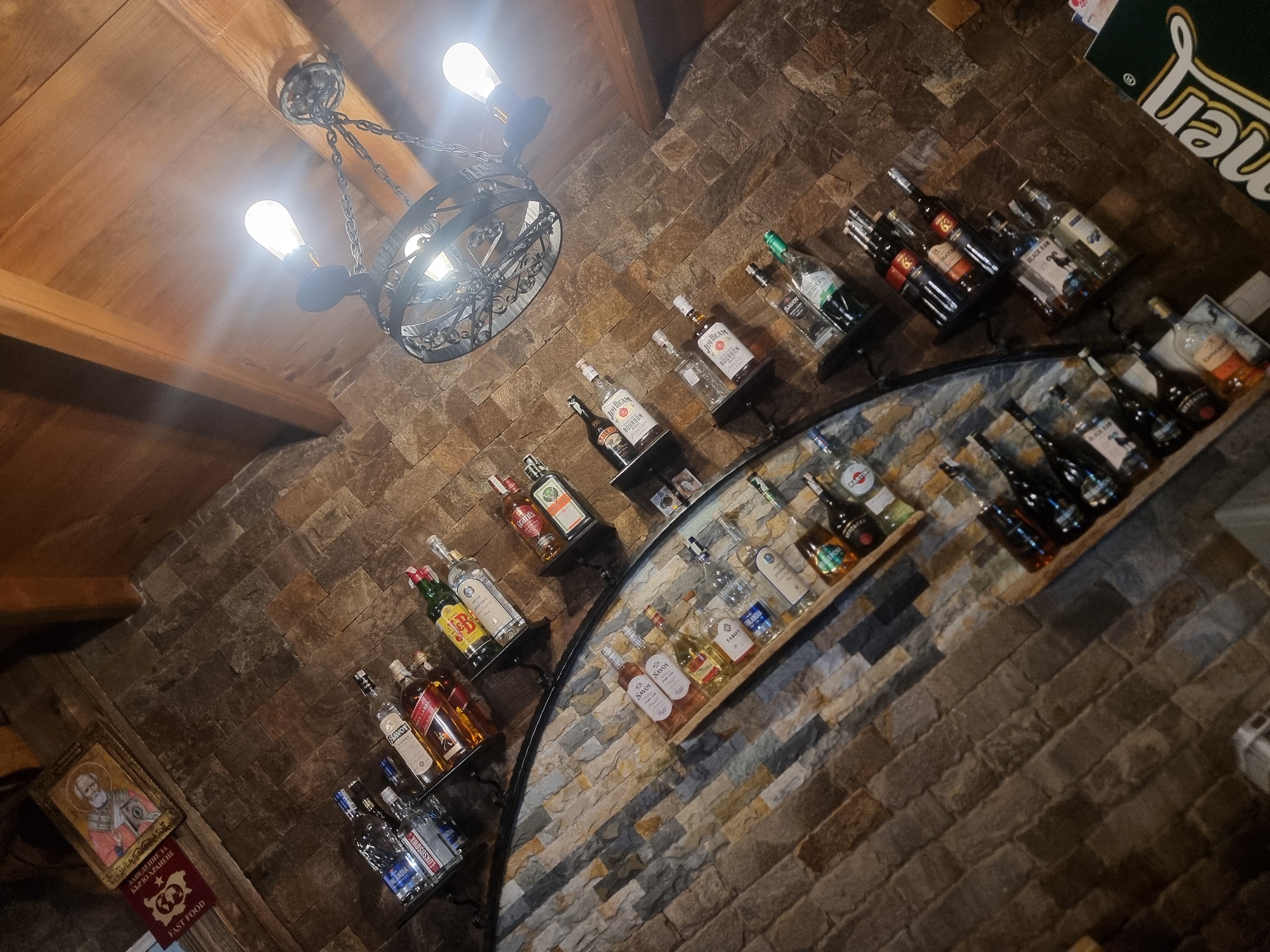 The image captures a bar or restaurant interior with a rustic aesthetic. The walls are constructed from irregularly shaped bricks of varying earth tones. A curved shelf system displays an assortment of liquor bottles, arranged along the wall in an arc. These bottles vary in shape, size, and label design, indicating a diverse selection of alcoholic beverages. A wrought iron chandelier hangs from the wooden ceiling, emitting a warm, bright light. On the bottom left, a framed icon and a sign with the words "FAST FOOD" in Cyrillic and English suggest the location might be in Banichan, Bulgaria. The perspective is tilted, giving a dynamic view of the scene.