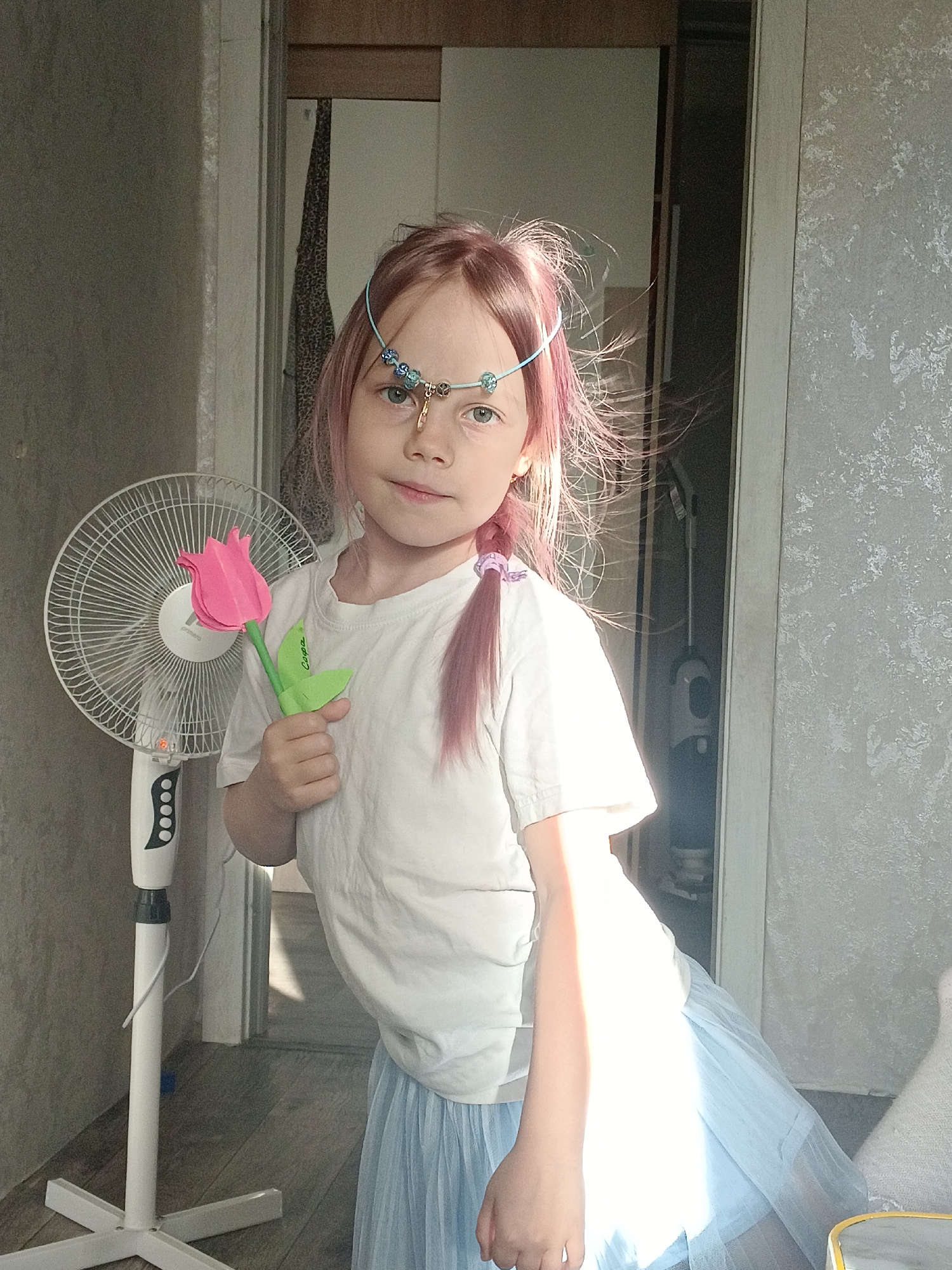 The image depicts a young girl with pink hair, adorned with a beaded headband, standing in an indoor setting. She is wearing a white t-shirt and a light blue skirt. In her right hand, she holds a pink paper tulip. To her left, a white oscillating fan is visible. The background shows a doorway leading to another room, with a floor made of wooden planks. The lighting suggests it is daytime, with sunlight casting shadows. The location is identified as Chaikovsky, Russia.