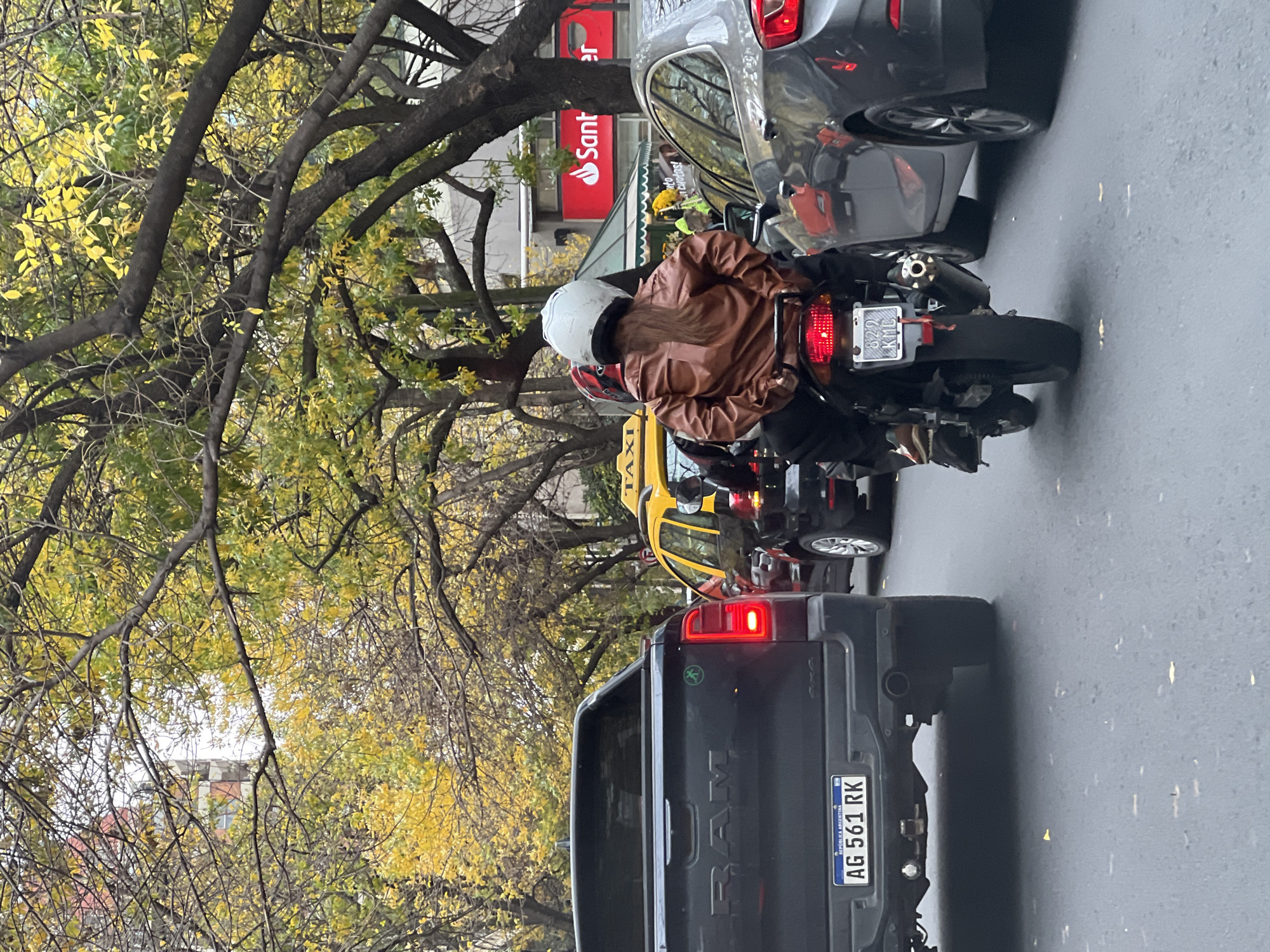 The image shows a street scene in Buenos Aires, Argentina. A black Ram pickup truck with license plate AG 561 RK is stopped in traffic. In front of it, a yellow taxi cab is also stopped. A motorcyclist wearing a brown jacket and a white helmet is on a motorcycle with a license plate that is difficult to read. The motorcycle is positioned between the taxi and a gray car, which is partially visible in the upper right corner. A Santander bank sign is visible in the background. Trees with yellow leaves overhang the street.