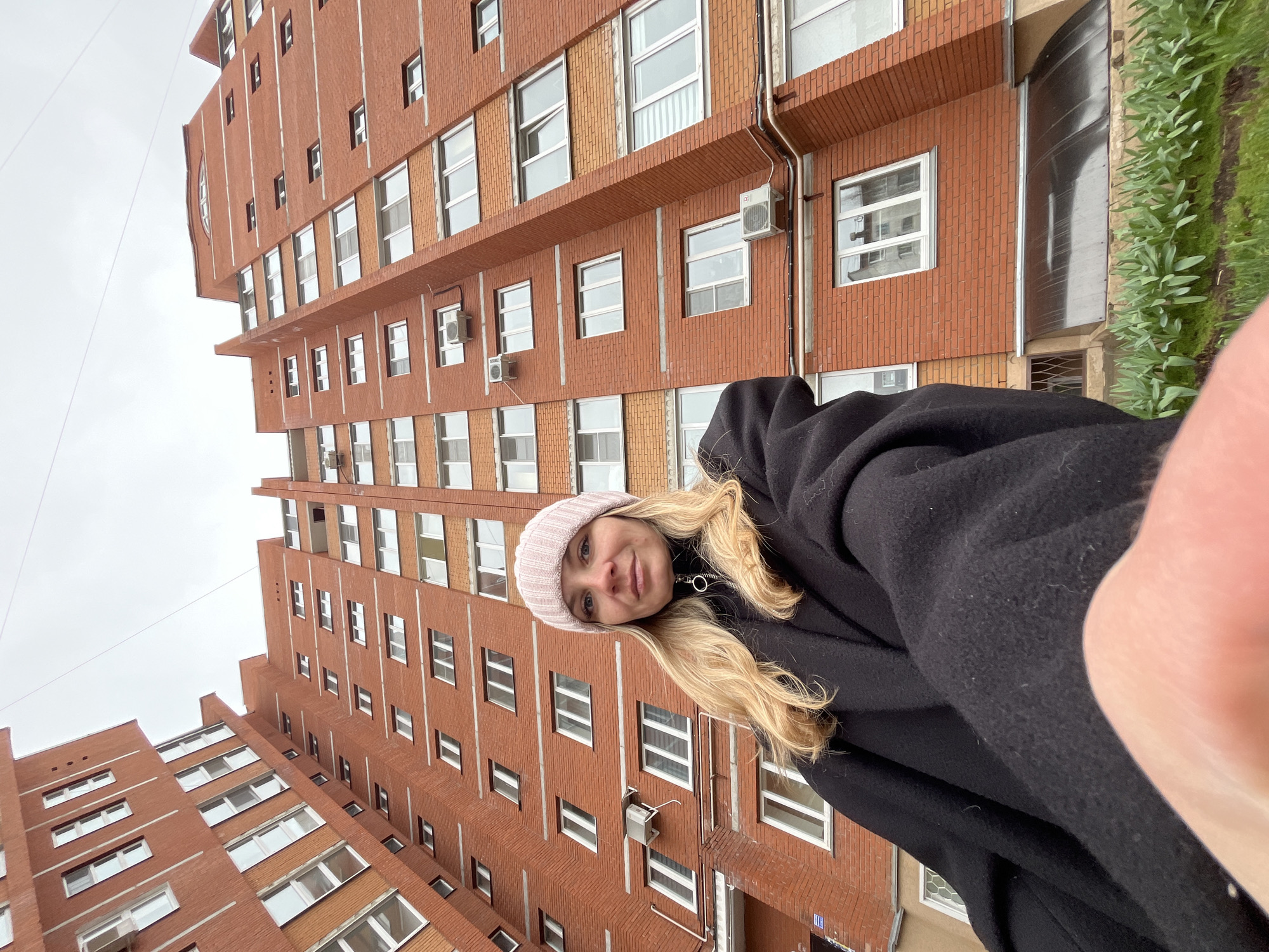 The media file depicts a young woman taking a selfie outdoors in Chaikovsky, Russia. She is positioned in the foreground, with a large, multi-story brick residential building filling the background. The woman has long, wavy blonde hair and is wearing a light pink knitted beanie and a black hooded coat or jacket. She is smiling faintly and looking directly at the camera, which she holds herself, with part of her hand visible in the lower right corner of the frame.
The building behind her is constructed of warm red brick, featuring numerous windows with white frames. Distinctive vertical white lines punctuate the brick facade, adding architectural detail. Several external air conditioning units are visible mounted below some of the windows. A narrow, brick-textured ledge or balcony is also present on one of the building's levels. To the far right, a strip of vibrant green foliage, possibly grass or small plants, grows alongside the building, suggesting ground level or a raised garden area. The sky above is uniformly light grey and overcast, indicating a cloudy daytime setting. A thin wire or cable stretches across the sky in the upper left quadrant. The perspective suggests the photo was taken from a low angle, looking upwards at both the woman and the tall structure. No visible text is present in the image.