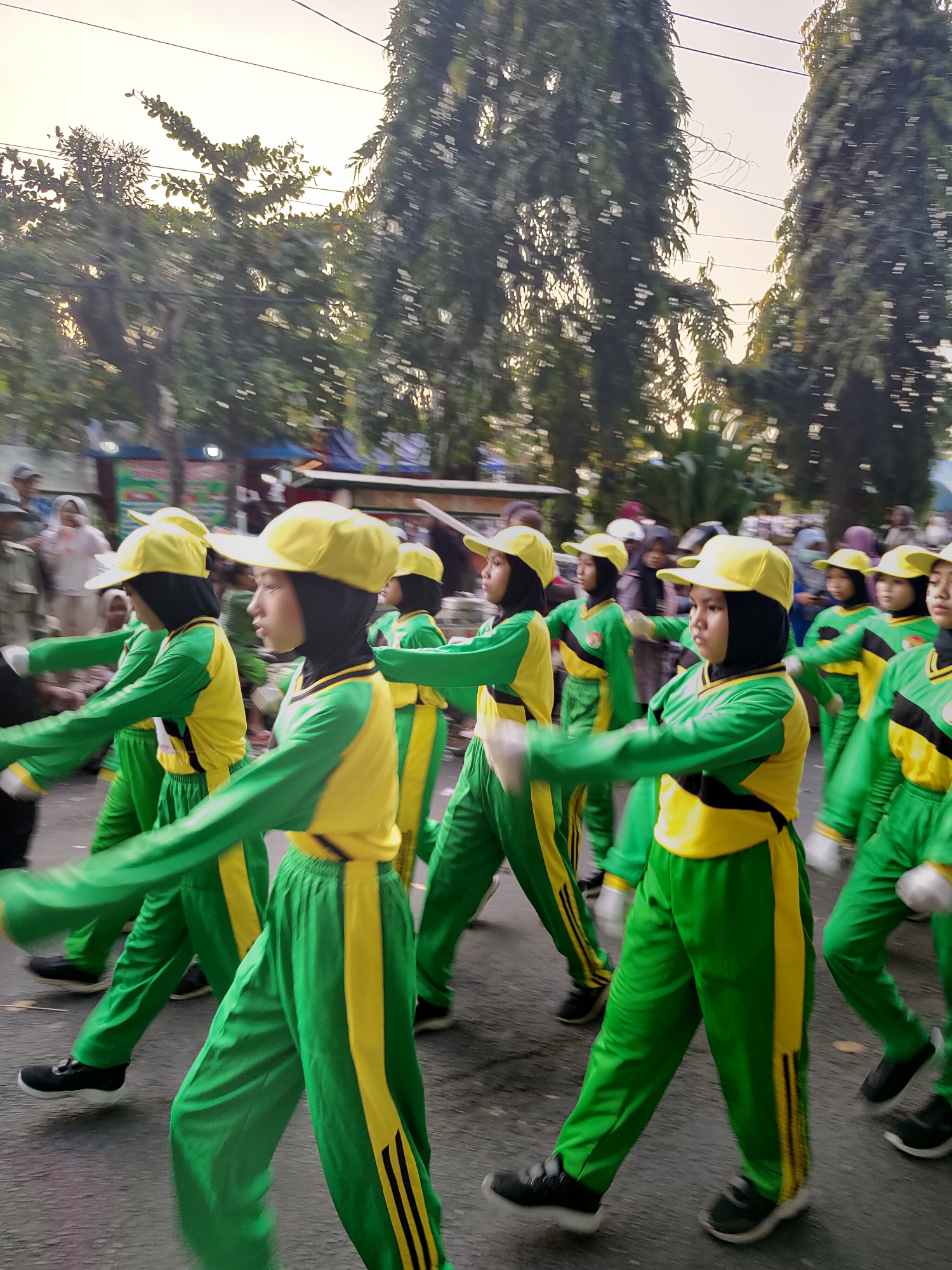 The image depicts a group of young people, likely students, participating in a parade or performance in Bangil, Indonesia. They are dressed in matching green and yellow uniforms with yellow caps and black hijabs. The uniforms feature long sleeves, matching pants, and what appear to be black shoes. Their arms are extended forward in a synchronized movement. The background is blurred, suggesting motion, and shows trees, some power lines, and hints of buildings and other people in the distance. The lighting suggests it could be early morning or late afternoon.