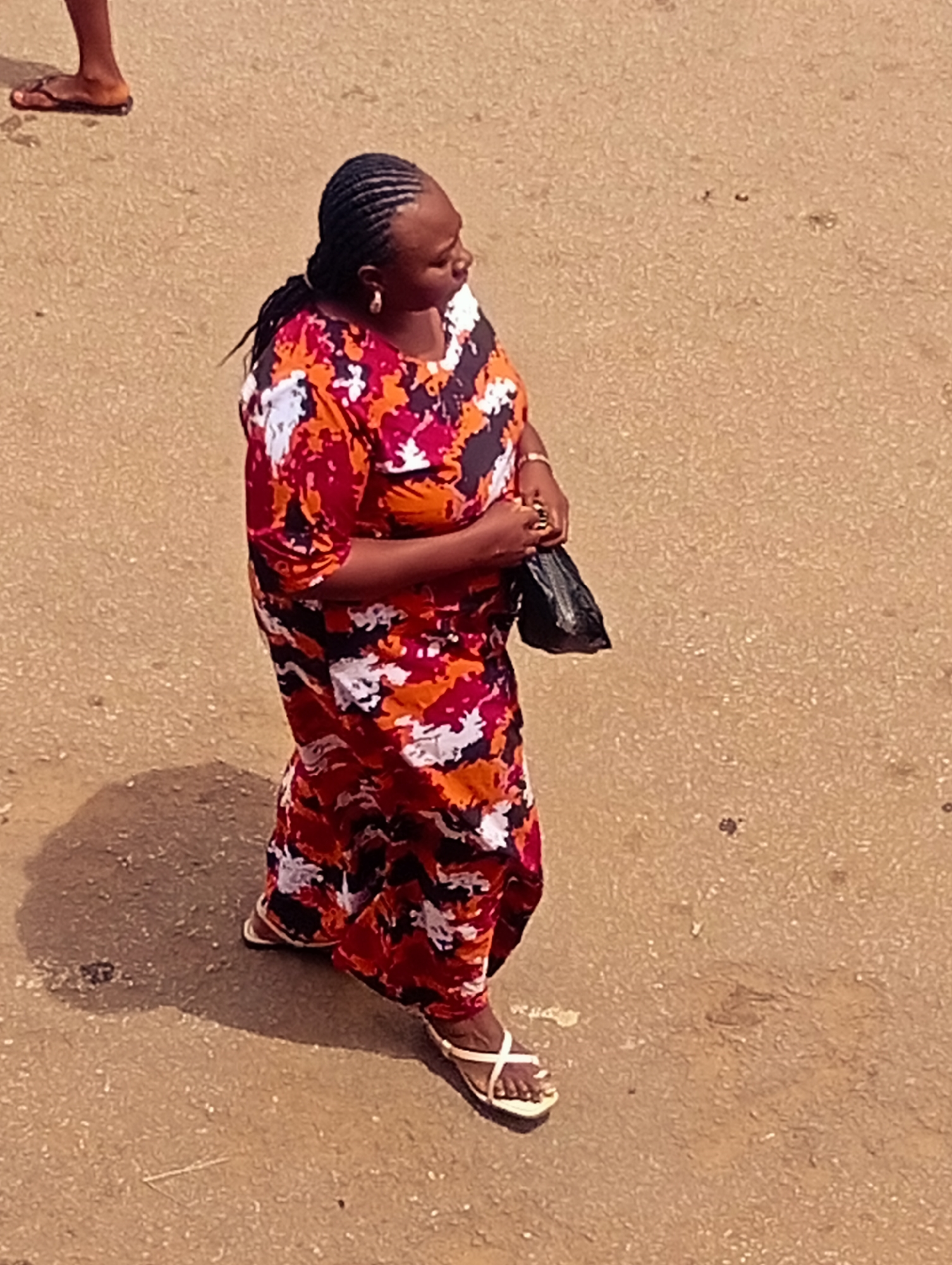 The image shows a woman walking on a paved surface, likely a road or a courtyard, in Bukuru, Nigeria. She is wearing a long, flowing dress with a colorful pattern of red, orange, white, and dark blue. The dress has short sleeves. On her feet, she wears white flip-flops with a strap across the toes. She is carrying a black plastic bag in her right hand. Her hair is styled in braids, and she is wearing hoop earrings.
To her left, the lower leg and foot of another person are visible, wearing brown flip-flops. The ground is a sandy, textured surface with some small stones and debris. There is a shadow cast on the ground to the woman's left, suggesting the sun is to her right and casting the shadow. The lighting indicates it is daytime, likely midday due to the defined shadow. The overall scene appears to be an outdoor setting in a somewhat dusty or unpaved area, consistent with many locations in Nigeria. There is no visible text in the image. The woman's posture suggests she is walking purposefully.