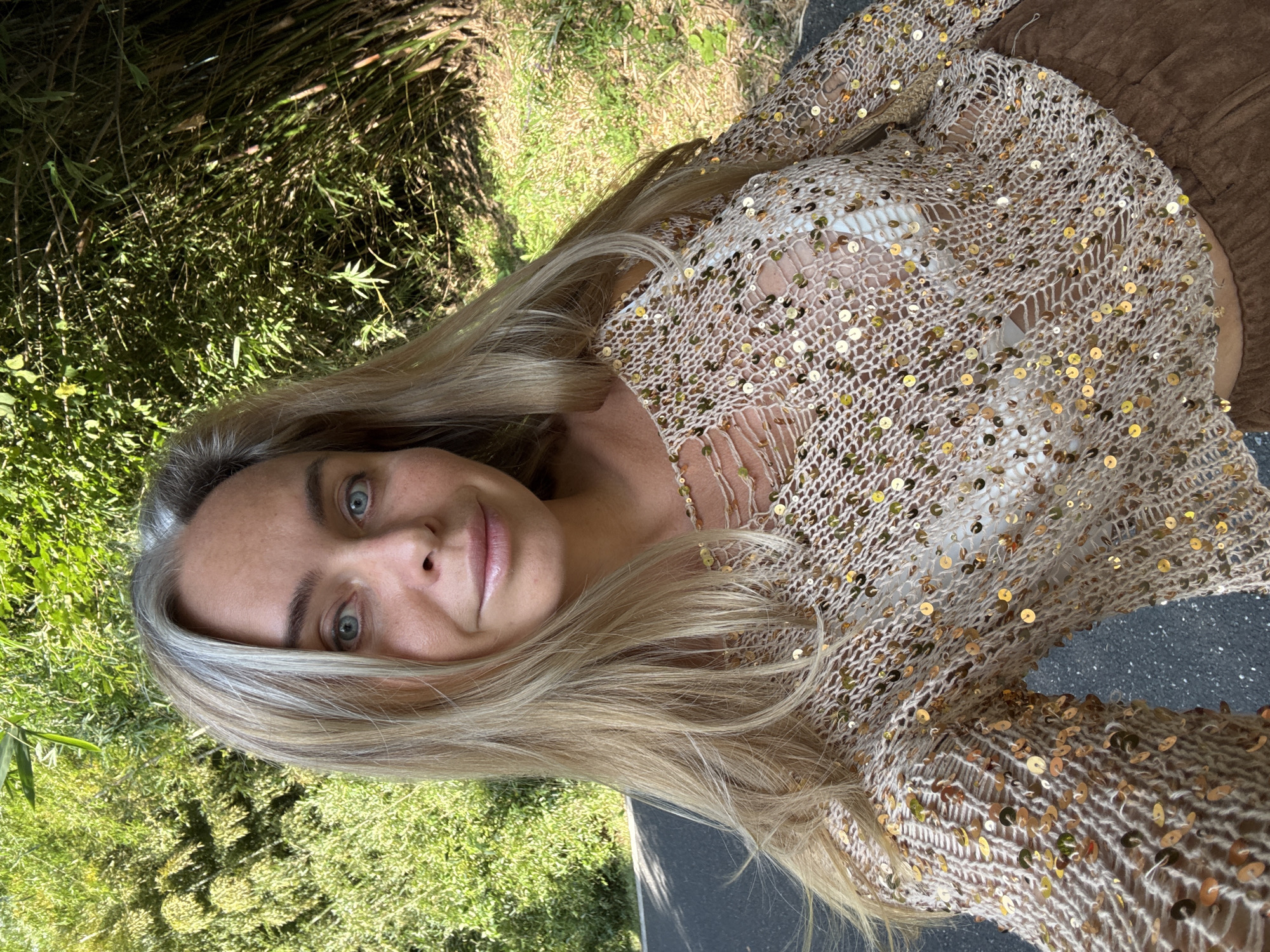 A woman with blonde hair and blue eyes is taking a selfie outdoors. She is wearing a light-colored, mesh top with gold sequins and a white bra underneath, and brown pants. She is standing or sitting on a paved surface, possibly a road or path. Behind her, there is lush green vegetation, including bamboo, and sunlight filters through the leaves, creating dappled shadows. The overall setting appears to be a natural, possibly park-like environment within the city of Wuxing District, China.