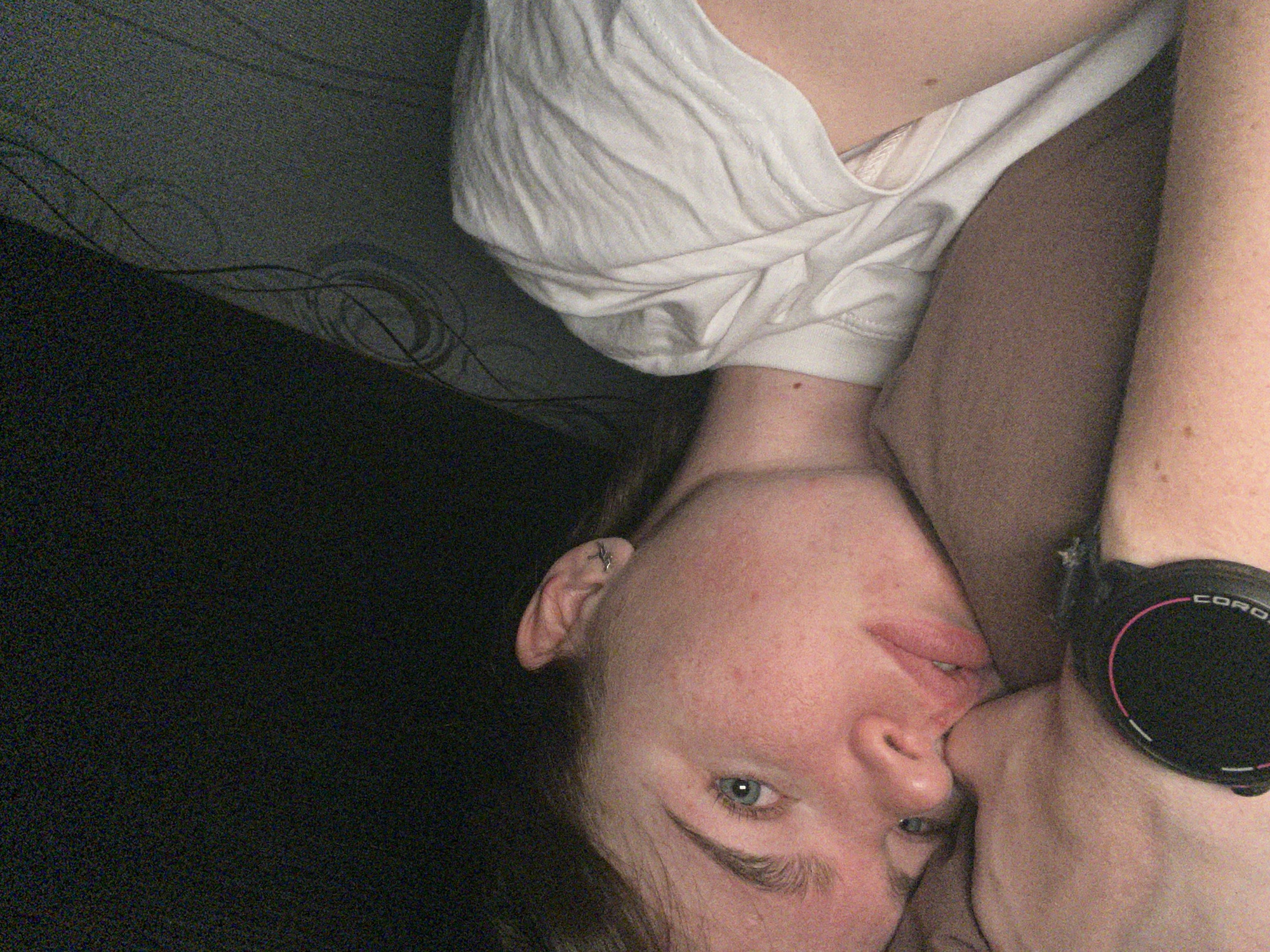A young adult female with light complexion, light eyes, and light brown hair is depicted in a dimly lit indoor setting in the city of Chaikovsky, Russia. She is lying on her left side, resting her head on her left arm, and looking directly at the camera with a neutral expression. Her face shows visible freckles or blemishes. A small metallic ear cuff is present on her right ear. She is wearing a white t-shirt. A black digital sports watch with a pink inner ring and "COROS" branding is visible on her left wrist. The immediate background behind her head is a dark brown fabric, while the upper right portion of the image reveals a dark patterned surface, possibly wallpaper or a textile, featuring swirling motifs. The upper left background is uniformly dark. The image exhibits significant grain due to low light conditions.