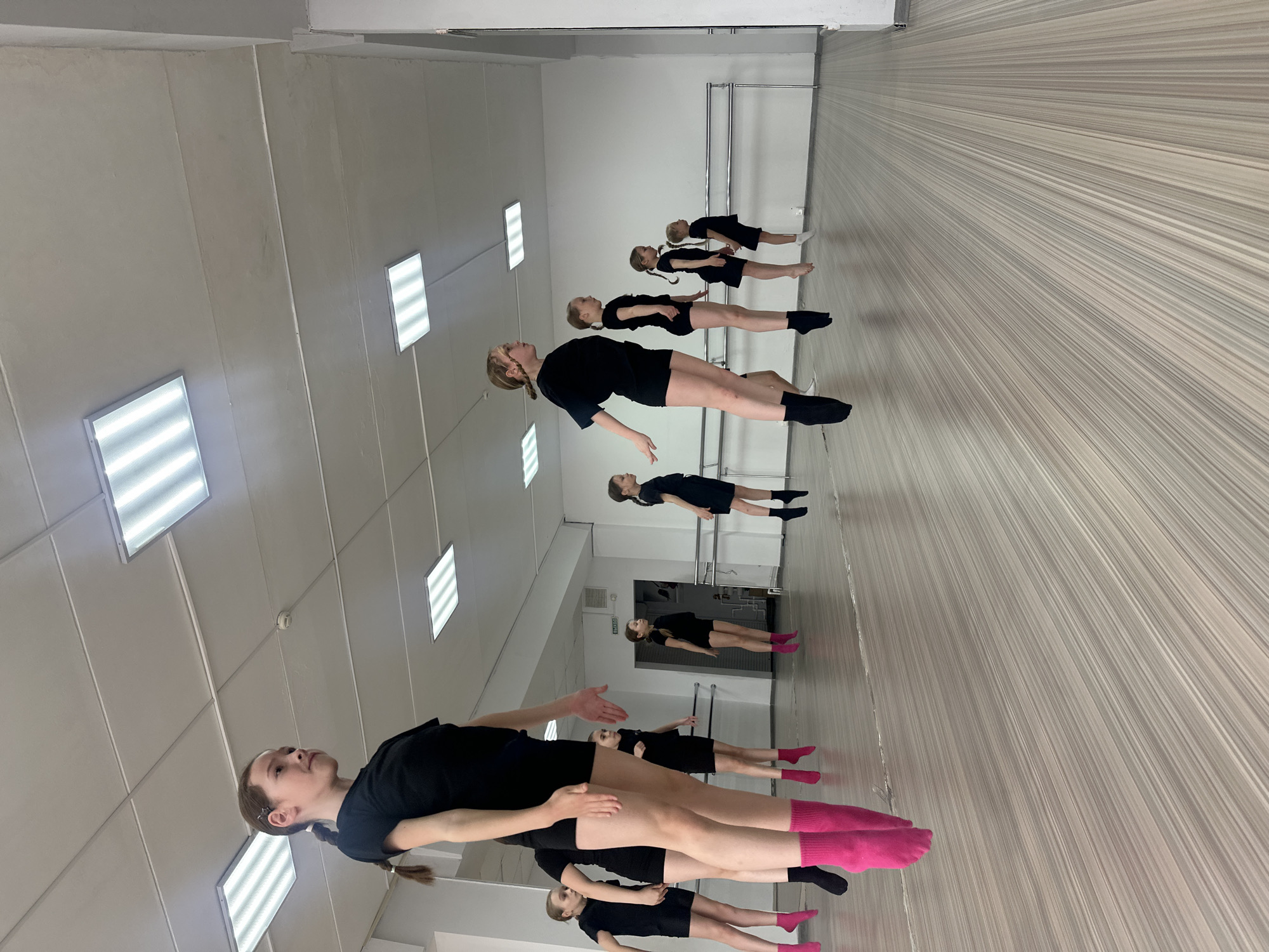 A group of five young females wearing black shirts and shorts are standing in a dance studio or similar space. The females are lined up in a row, facing forward. They are wearing various colored socks. The studio has a wooden floor with parallel lines. On the wall is a mirrored reflection of the girls and space. The location is a dance studio or similar space in Votkinsk, Russia.
