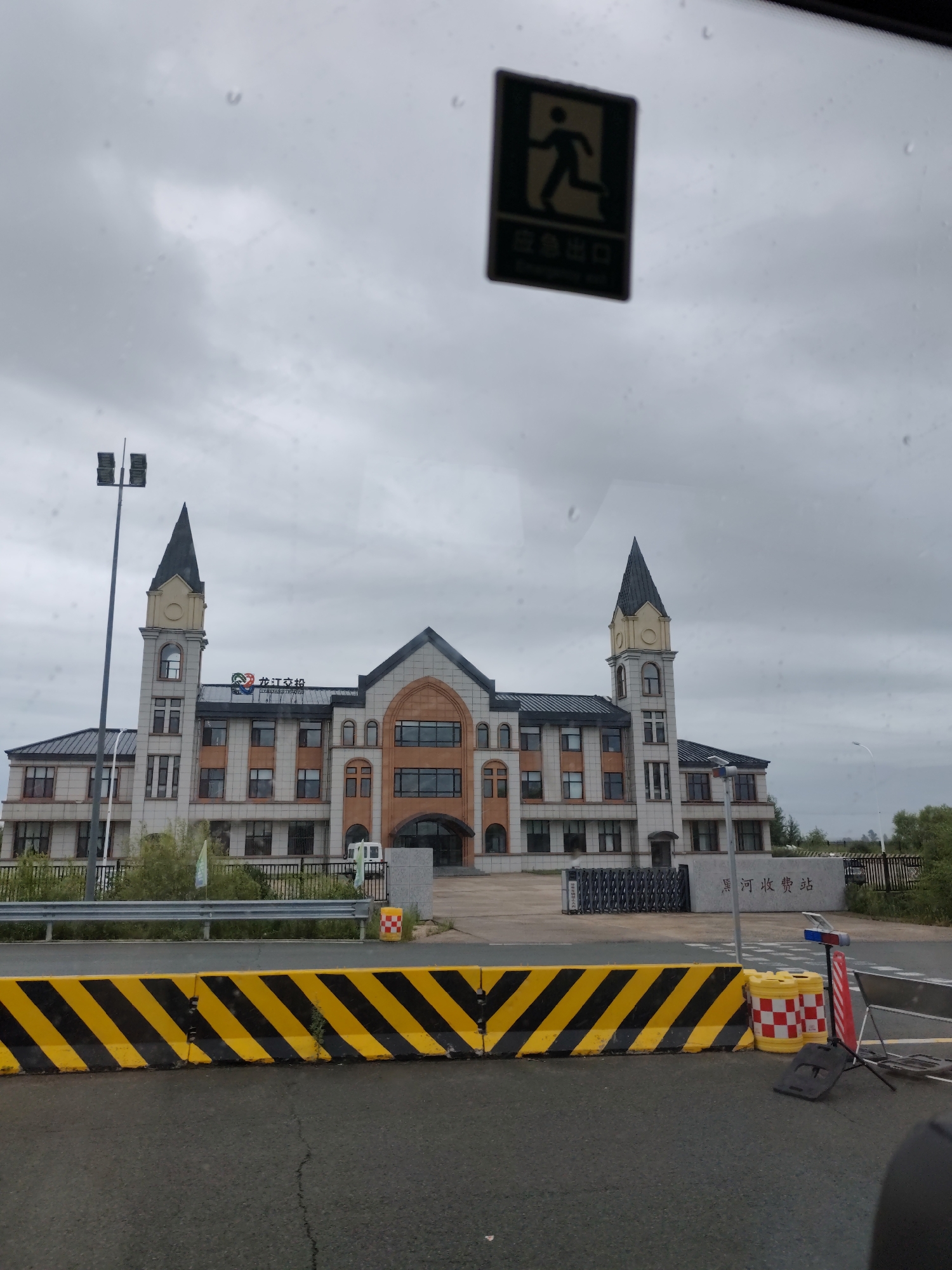 The image shows a building with two towers, a light pole, and a sign in the city of Aihui, China. The building appears to be a government or administrative building, with a modern architectural style incorporating some classic elements. The sign in front of the building reads "Heihe Toll Station" in Chinese characters, indicating its location and function. The sky is overcast, suggesting a cloudy or rainy day. The foreground is occupied by a road with a yellow and black striped barrier, likely a traffic control measure. There are no people or animals clearly visible in the image. The overall scene depicts a functional infrastructure area within an urban or semi-urban setting.