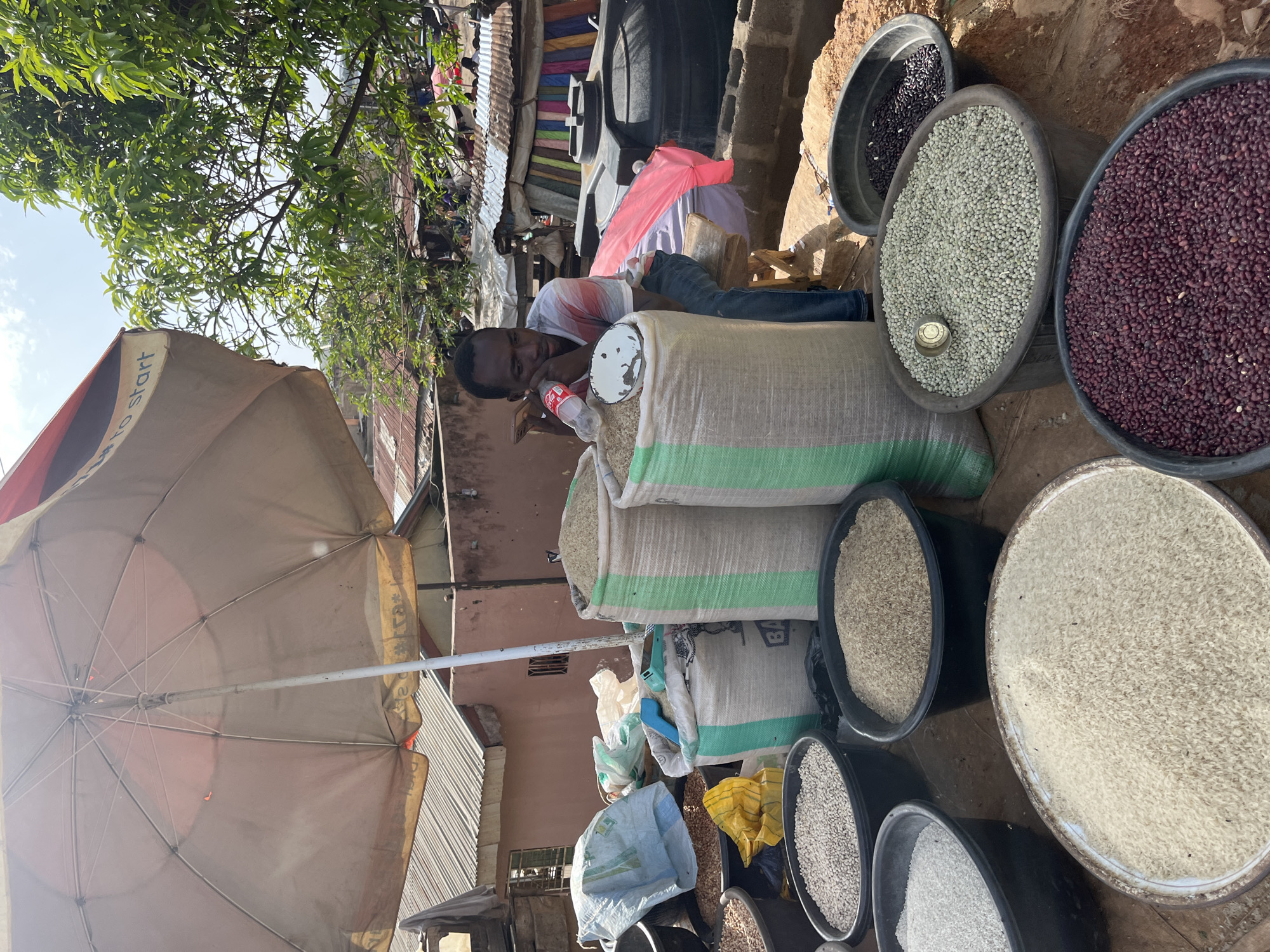 This media file captures a market scene in Jos, Nigeria, likely in the daytime given the bright sunlight. The image is oriented vertically, with the sky and trees visible at the top, suggesting an outdoor setting. The central subject is a young Black man, lying down and looking towards the camera, holding a red and white bottle of what appears to be Coca-Cola. He is wearing a striped t-shirt and blue jeans.
The man is positioned near large sacks of grain, some with green and white stripes, and several large black basins filled with various types of grains and beans. Visible in the basins are what appear to be black beans, light green peas, and dark red beans, along with different varieties of rice. A pale beige umbrella, partially unfurled, provides shade over a portion of the market stall. In the background, there are corrugated metal roofs, mud brick walls, and colorful fabrics, indicating a busy, possibly informal, marketplace. There are also plastic water tanks and bags, adding to the utilitarian atmosphere of the setting.
The man's expression is neutral, perhaps a bit tired or relaxed. The overall mood is that of everyday life in a Nigerian market, with a focus on the agricultural products being sold. There is some visible text on the umbrella, appearing to be in English, but it is mostly obscured and difficult to read clearly. The sunlight casting shadows and highlighting the textures of the grains and objects suggests it is likely late morning or early afternoon.