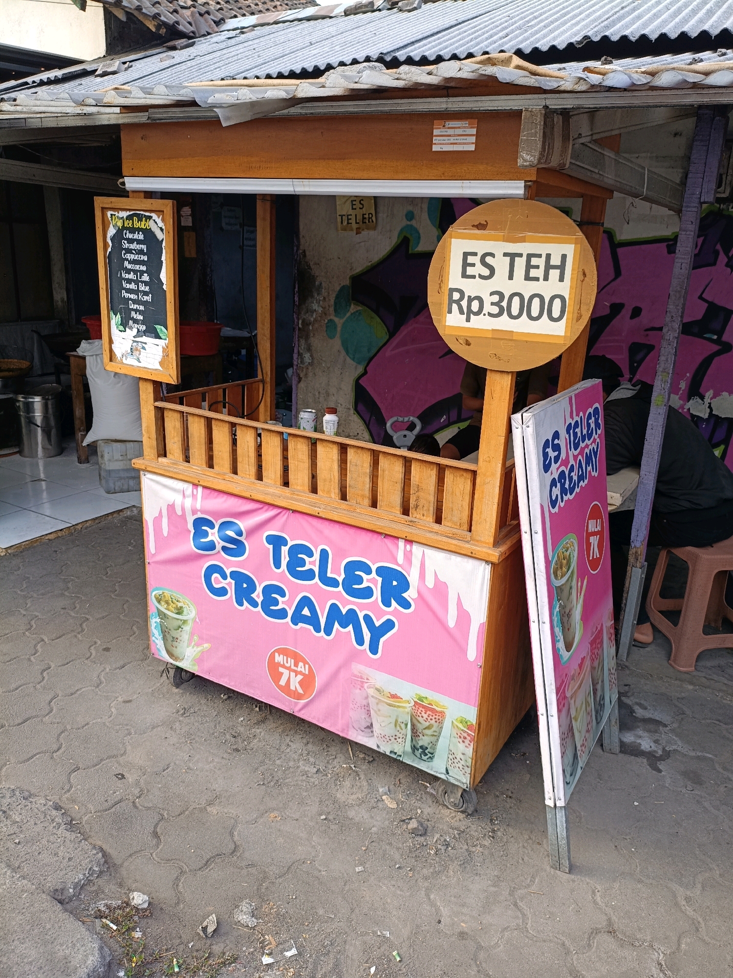 The image shows an outdoor food stall in Maguwoharjo, Indonesia, selling "Es Teler Creamy". The stall has a wooden structure with a pink tarp displaying the name of the product and images of drinks. A separate sign displays "ES TEH Rp.3000". There is a menu board on the left with a list of drinks. The stall is set up on a paved area with some debris. In the background, there is a building with a corrugated metal roof and graffiti on the wall. A person wearing a dark jacket is partially visible in the background, seated on a stool.