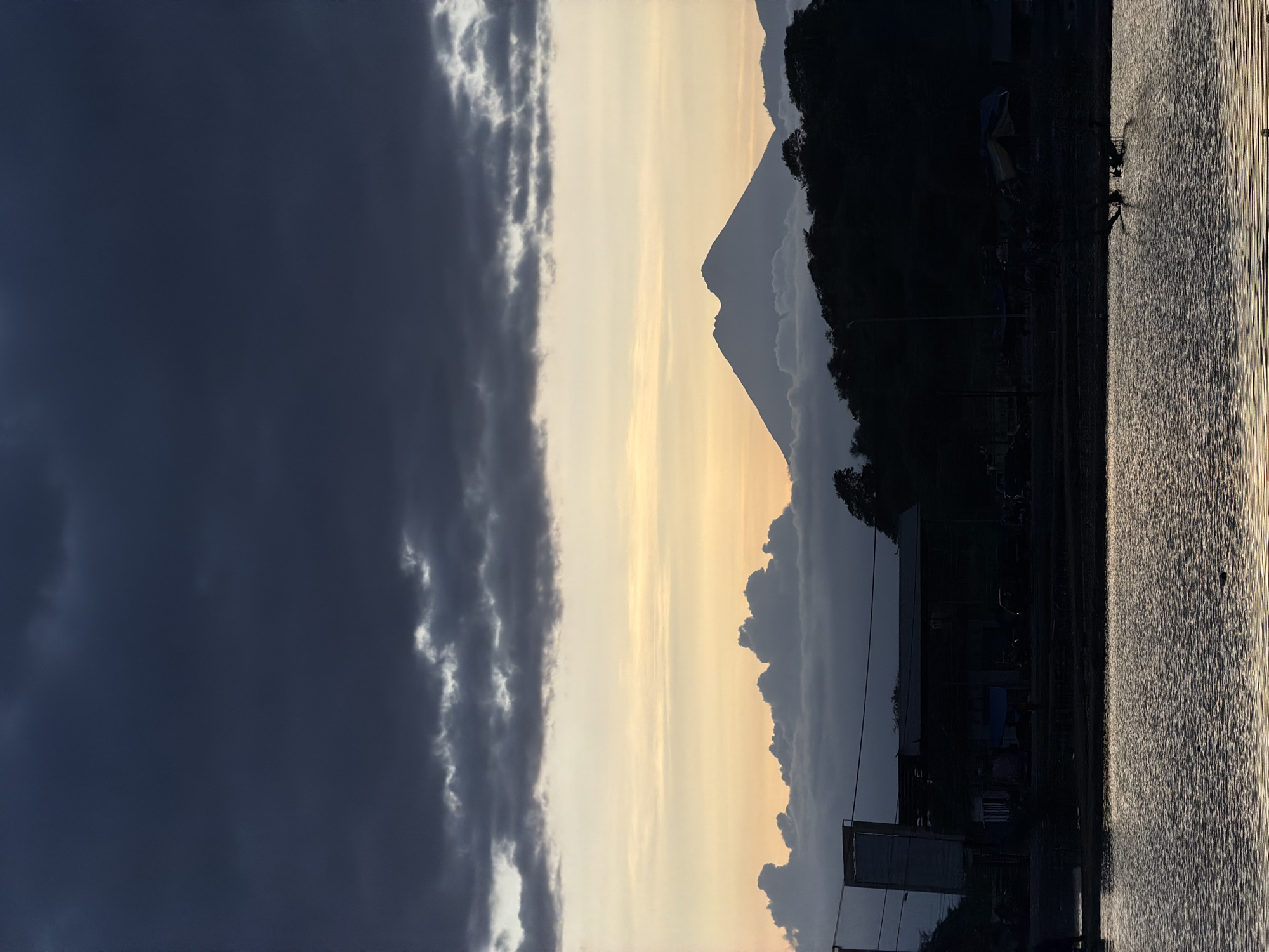 The image depicts a landscape at dusk or dawn, possibly located near Upt Tambak Sari, Indonesia. The sky is divided between a dark, stormy area on the left and a lighter, yellowish sky on the right. A silhouetted mountain range is visible in the center, with a distinct pointed peak. A dark, undefined area, potentially a body of water, occupies the right side of the image, reflecting the light. On the horizon, silhouetted buildings and structures are visible. The image is rotated 90 degrees counterclockwise.
