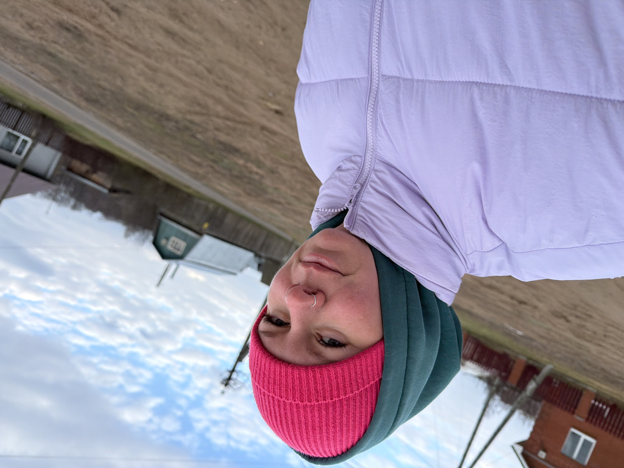 The image shows a close-up portrait of a person, likely a woman, taken outdoors against a blurred rural or semi-rural background. The person is looking directly at the camera from a low angle, suggesting a selfie.
**Subjects:** The main subject is a person wearing a bright pink knitted beanie, a dark green hoodie, and a light purple puffer jacket. The person has a silver nose ring on their left nostril and appears to have a neutral to slightly content expression. Their skin has a natural, slightly flushed tone, possibly due to the cool outdoor temperature.
**Scene and Setting:** The background features a partly cloudy sky at the top. Below the horizon line, there's a stretch of brown, dry ground, possibly a field or roadside, with a narrow path or road running through it. In the mid-distance, several buildings are visible, including a reddish-brown house with a white window and a light blue or green building. Fences and utility poles or bare trees are also present, suggesting a non-urban, possibly rural or suburban environment. Given the context of Chaikovsky, Russia, this fits a description of a countryside or small town setting.
**Activity or Event:** The image captures a moment of the person posing for a photo, likely a casual outdoor selfie.
**Notable Details:** The time of day appears to be daytime, with a bright, overcast sky. The warm winter clothing (puffer jacket, hoodie, beanie) suggests cool or cold weather, aligning with late autumn, winter, or early spring in a northern climate. The strong contrast in colors between the bright pink beanie, dark green hoodie, and light purple jacket is prominent. No visible text is present in the image.