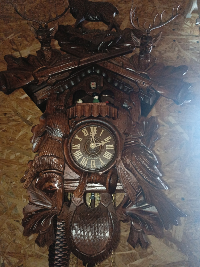 In Gorka, Russia, a large, ornate wooden cuckoo clock, featuring a dark brown finish and intricate carvings, is affixed to a light-colored, pressed wood panel wall. The clock's uppermost section displays a carved deer stag standing atop a platform of foliage. Below this, two carved deer heads with prominent antlers flank a pair of crossed hunting rifles or horns. Directly beneath the crossed implements, a small balcony houses two small, articulated figures: one predominantly white with dark spots, and another with a green body and black hat, positioned next to a small red object. Below the balcony, the central circular clock face features white Roman numerals, gold-toned hands, and the inscription "MADE IN GERMANY." The time displayed is approximately 1:55. Flanking the clock face are large, deeply carved predatory birds, with additional foliage and smaller bird carvings on the left side. A carved hunting horn is positioned horizontally beneath the clock face. The lower section of the clock is adorned with a large, textured carved shield or pouch, with a carved pine cone weight suspended by a chain visible below it. Another chain is partially visible to the right of the shield. The overall scene is illuminated with a strong light source originating from the right, causing a bright glare.