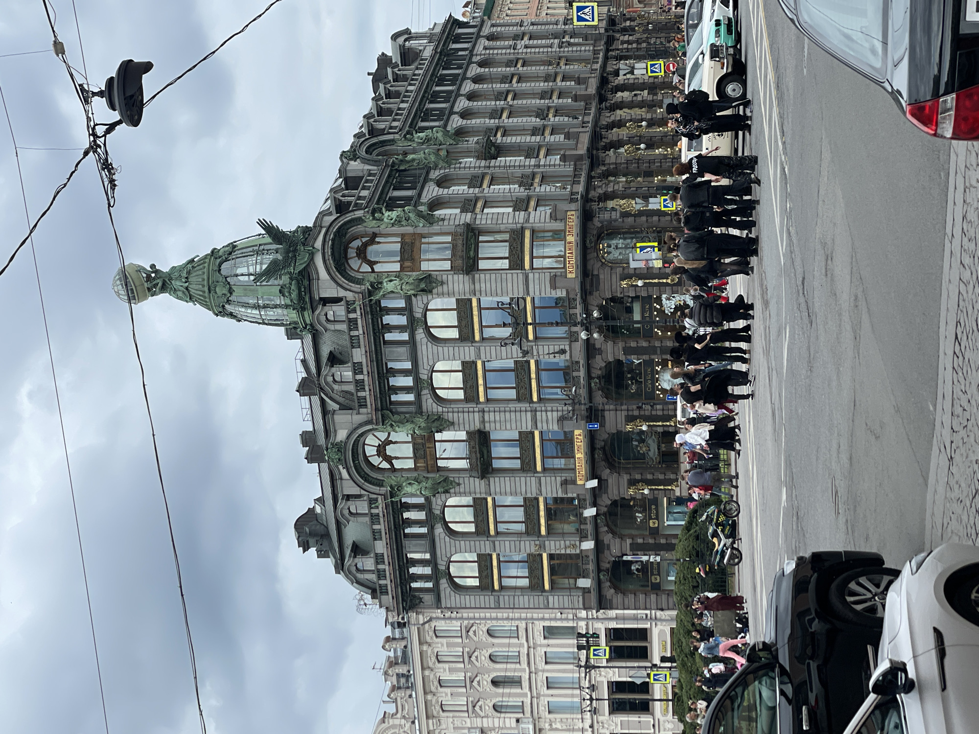 This is a full-shot, eye-level, outdoor, daytime photograph of a street scene in Saint Petersburg, Russia, featuring the Singer House building. The Singer House, also known as the Dom Knigi (House of Books), is a prominent Art Nouveau building located at Nevsky Prospekt 28.
The central subject is the Singer House, a grand, multi-story building with a distinctive green dome topped by a glass globe and an eagle sculpture. The facade is adorned with numerous arched windows, decorative sculptures of winged figures, and intricate architectural details. The building's exterior is primarily gray stone with gold accents. The ground floor appears to house retail spaces, with signage visible, including "STORE" and "Cafe Rene".
In the foreground, a gray paved street with white lane markings is visible. Several cars are parked or driving on the street, including a black SUV and a white car in the lower left portion of the frame, and a white taxi with green and turquoise accents on the right.
A group of approximately 15-20 people are gathered on the sidewalk and street in front of the Singer House, some appearing to be posing for photos or observing the building. They are dressed in casual, modern clothing. Among them, a person in a white hoodie stands out. A motorcycle is parked on the sidewalk near the group.
Pedestrian crossing signals and street signs are present, indicating an active intersection. The sky is overcast with heavy gray clouds, suggesting a cool or potentially rainy day. The overall atmosphere is that of a busy urban environment with tourists and locals.