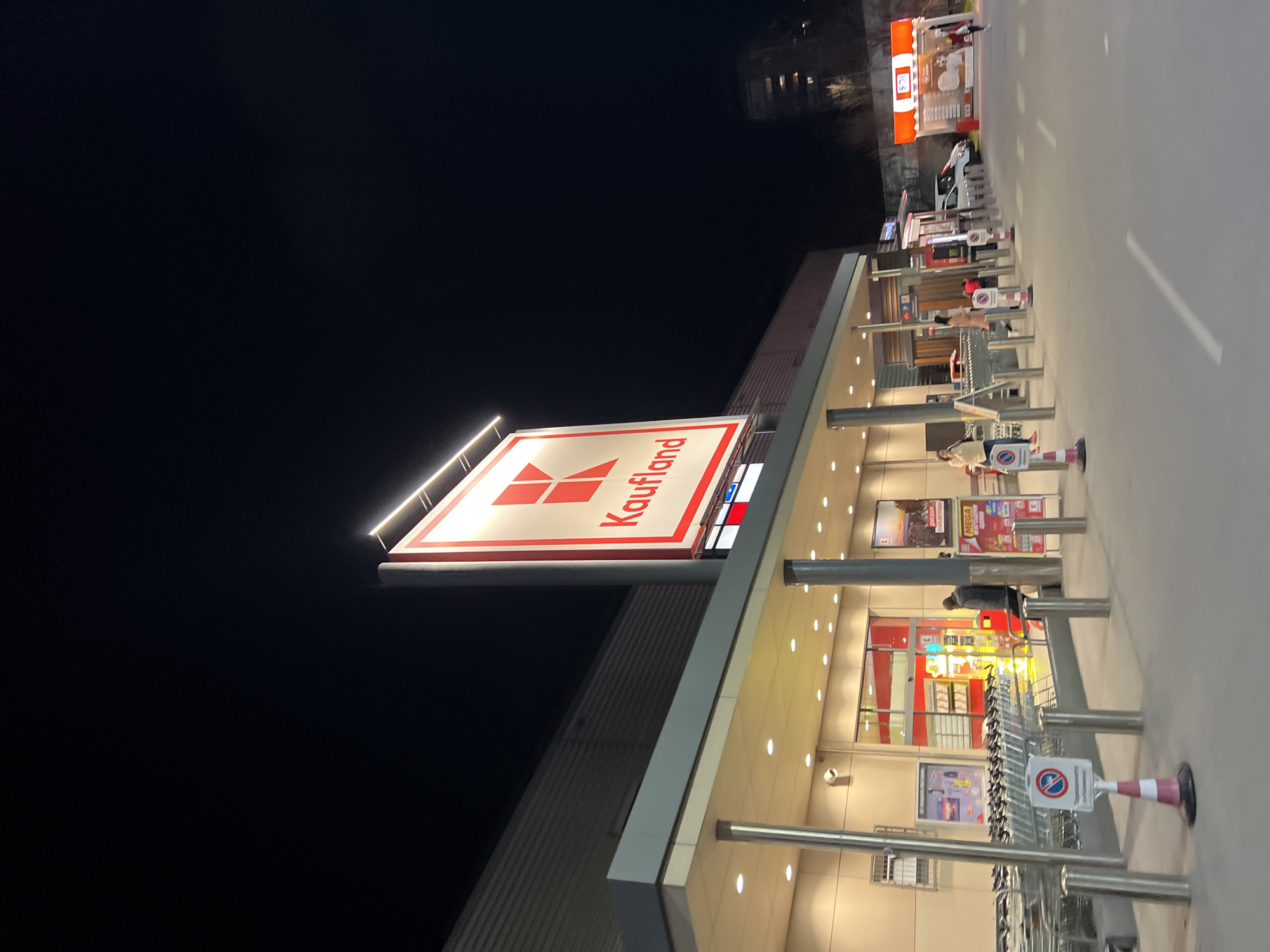 The image depicts the exterior of a Kaufland supermarket in Plovdiv, Bulgaria, at night. The large, illuminated Kaufland sign is prominently displayed. The building facade is visible, along with rows of shopping carts. There are a few people present near the entrance. In the background, there is a building and what appears to be a bus station.