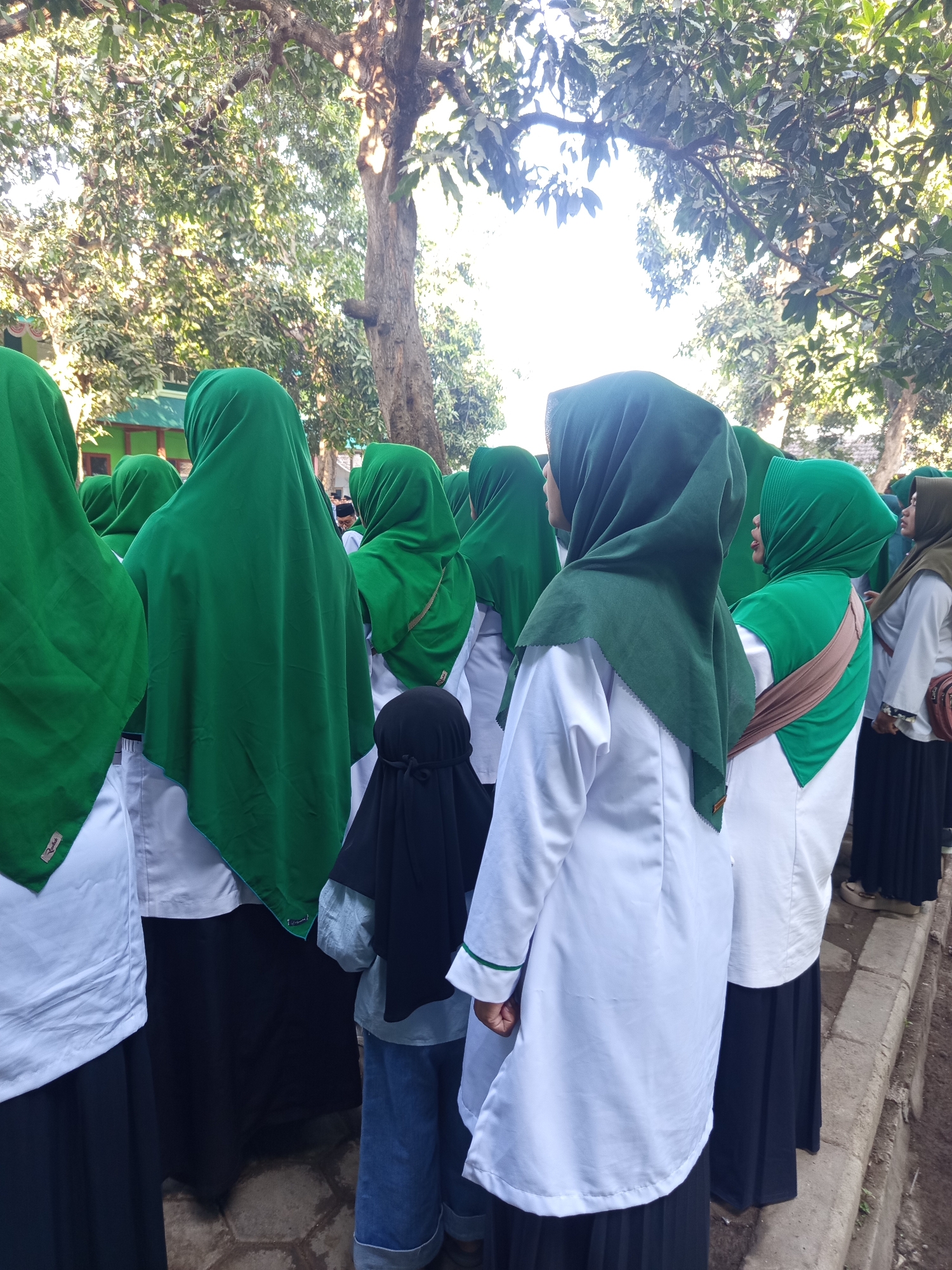 In Lumutan, Indonesia, a group of young girls in uniform are seen from the back, standing outdoors. They are all wearing white shirts and dark skirts, with the majority of them sporting vibrant green hijabs. One girl in the foreground has a dark black niqab that covers her face, leaving only her eyes visible, and she is wearing blue jeans. The setting appears to be near a building with a green roof, surrounded by lush trees. The ground beneath them is paved with interlocking stones. The overall scene captures a moment of communal gathering or activity among these students in the Indonesian town.