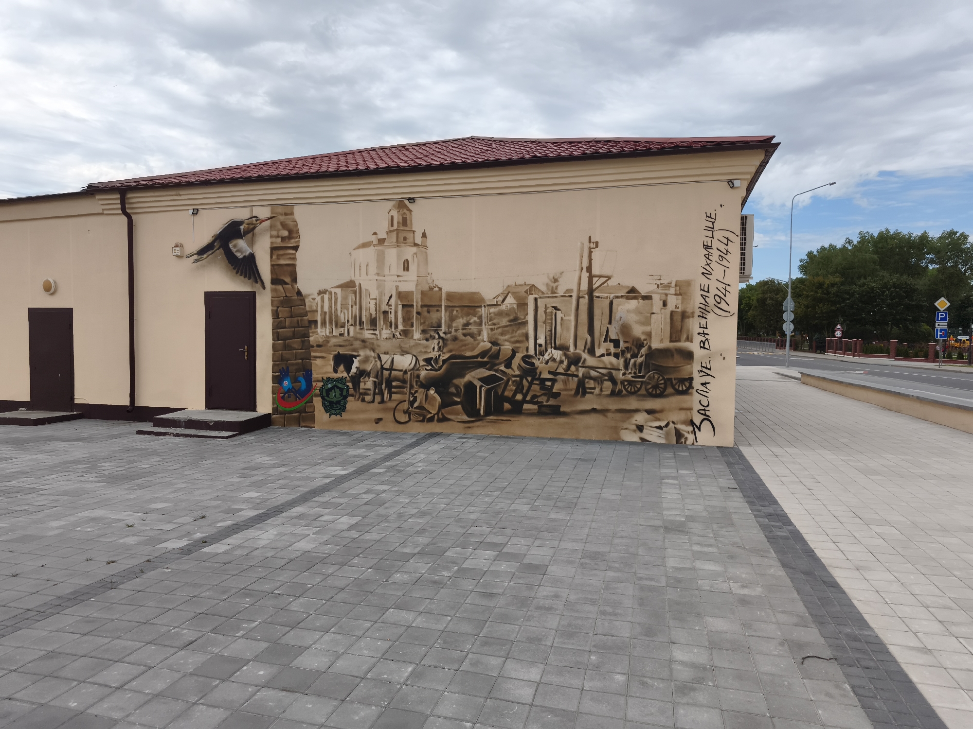 The image depicts a building with a large mural on its side, located in the city of Zaslavl, Belarus. The mural is monochromatic and appears to be rendered in shades of brown and white, depicting a historical scene. In the foreground, a bird is shown in flight. The central part of the mural features a town square with a church, horse-drawn carts, and people engaged in various activities, suggesting a historical marketplace or event. The style is detailed and realistic, evoking a sense of the past. On the right side of the mural, text in Cyrillic script reads "ЗАСЛАУЕ. ВАЕННАЕ ПАМЯЦЕЛЕ. (1941-1944)", which translates to "Zaslavl. Military Memorial. (1941-1944)", indicating the mural commemorates events during World War II in this location.
The building itself is modern, with a light-colored facade and a red tiled roof. Two dark-colored doors are visible on the left side of the building, with concrete steps leading up to them. Adjacent to the mural, on the far left of the wall, there is a smaller, more colorful crest or emblem, possibly representing a local symbol.
The area in front of the building is paved with grey interlocking stone tiles, laid out in a uniform pattern. The lighting suggests it is daytime, with the sun casting shadows across the pavement. In the background, a street with traffic signs, a street lamp, and a line of trees can be seen. The sky is partially cloudy. The overall impression is one of a well-maintained public space featuring historical art.