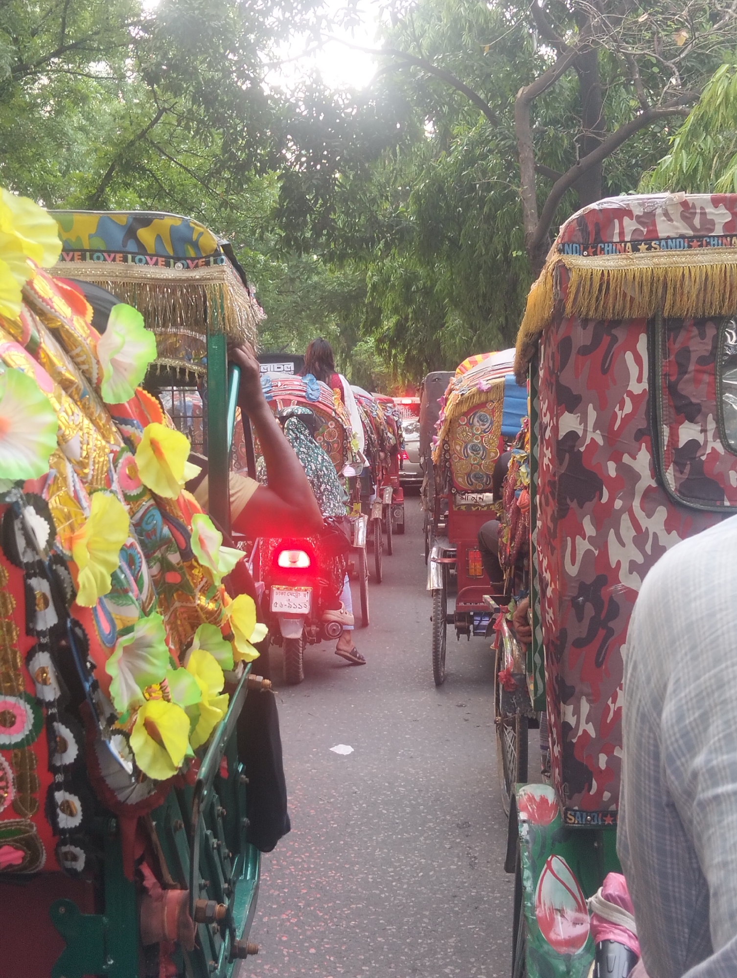 The image depicts a street in Dhaka, Bangladesh, congested with cycle rickshaws and auto rickshaws. The rickshaws are adorned with colorful decorations and embellishments. The photograph is taken from the perspective of someone within the traffic, showing a row of rickshaws and auto rickshaws ahead. Drivers and passengers are visible within the vehicles. The road surface is asphalt. Overhead, trees create a canopy, partially obscuring the sky. A motor vehicle is behind the closest cycle rickshaw with the driver's leg clearly showing and their brake lights on. The environment suggests a busy urban setting.