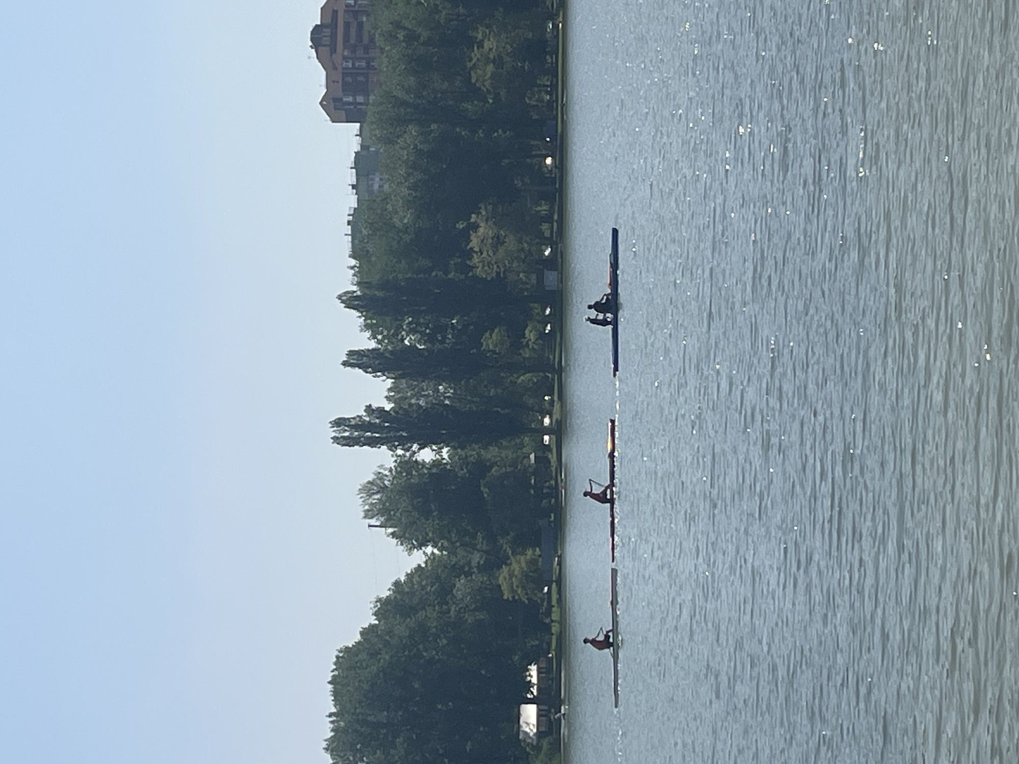 The image shows a body of water, likely a lake or wide river, with three rowing boats in the distance. The water has visible ripples and reflections. The shoreline is lined with dense trees and foliage. In the background, a building is visible, possibly an apartment complex. The scene appears to be in a city environment, specifically Krasnodar, Russia, based on location clues.
