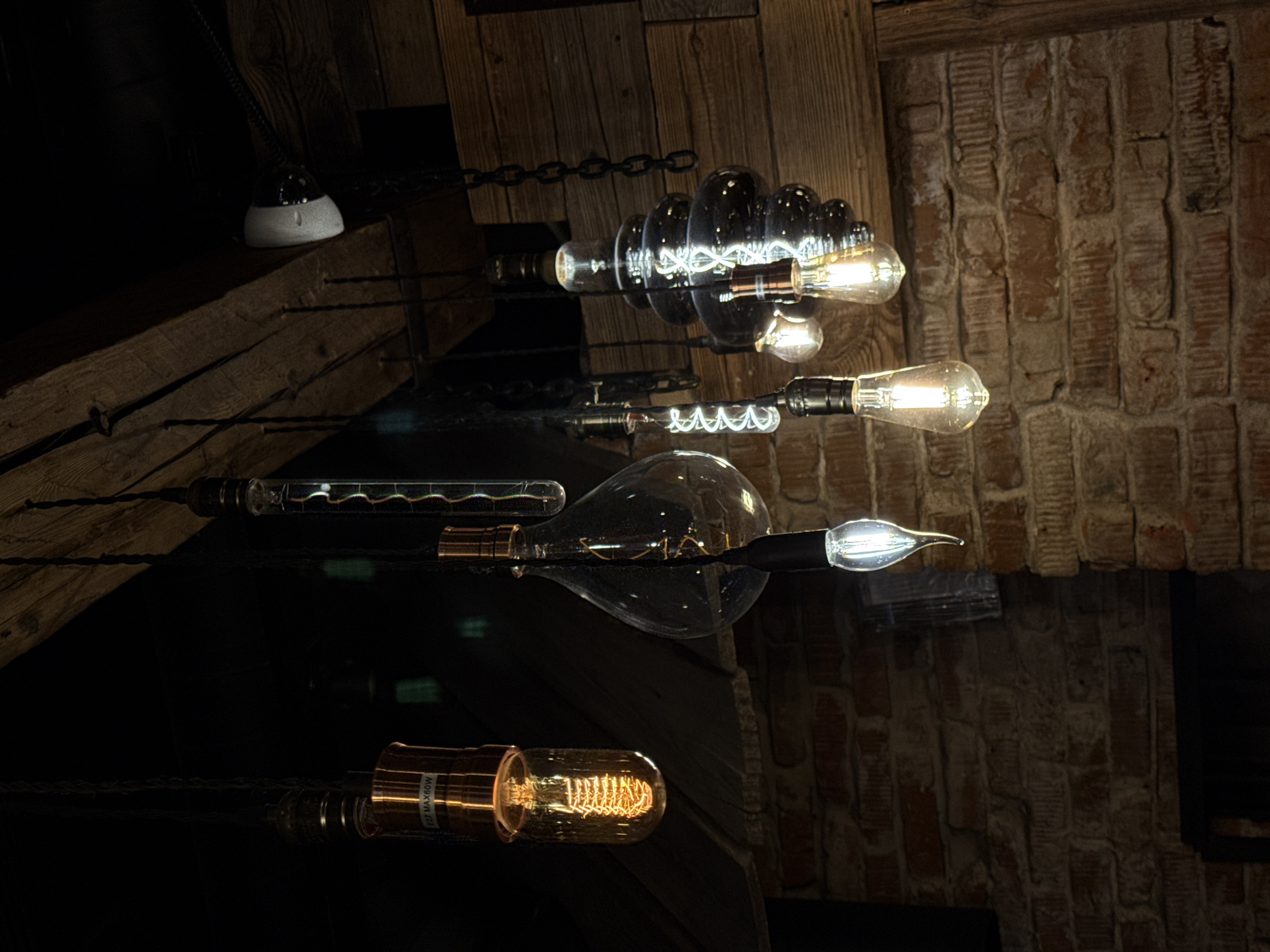 The image depicts a close-up, low-angle shot of an eclectic arrangement of vintage-style Edison light bulbs, hanging from dark cables and chains against a dimly lit, rustic interior. The background consists of rough brick walls and wooden beams, suggesting a restaurant or bar setting. The bulbs themselves are varied in shape and design, with some featuring exposed filaments that emit a warm, orange glow, while others are clear glass revealing intricate wire designs or are more modern LED equivalents. One bulb, hanging to the left, has a copper-colored socket and text that reads "E27 MAX60W". Another bulb, to the right, is a dark, multi-tiered glass structure, with a filament visible within. A single, modern-looking white bulb with a pointed tip hangs lower on the right. The lighting is dramatic, with the glow of the bulbs illuminating the immediate surroundings and casting shadows. There are no people or animals visible in the image. The overall impression is one of industrial-chic decor. The location of this scene is unknown and cannot be determined from the provided image.