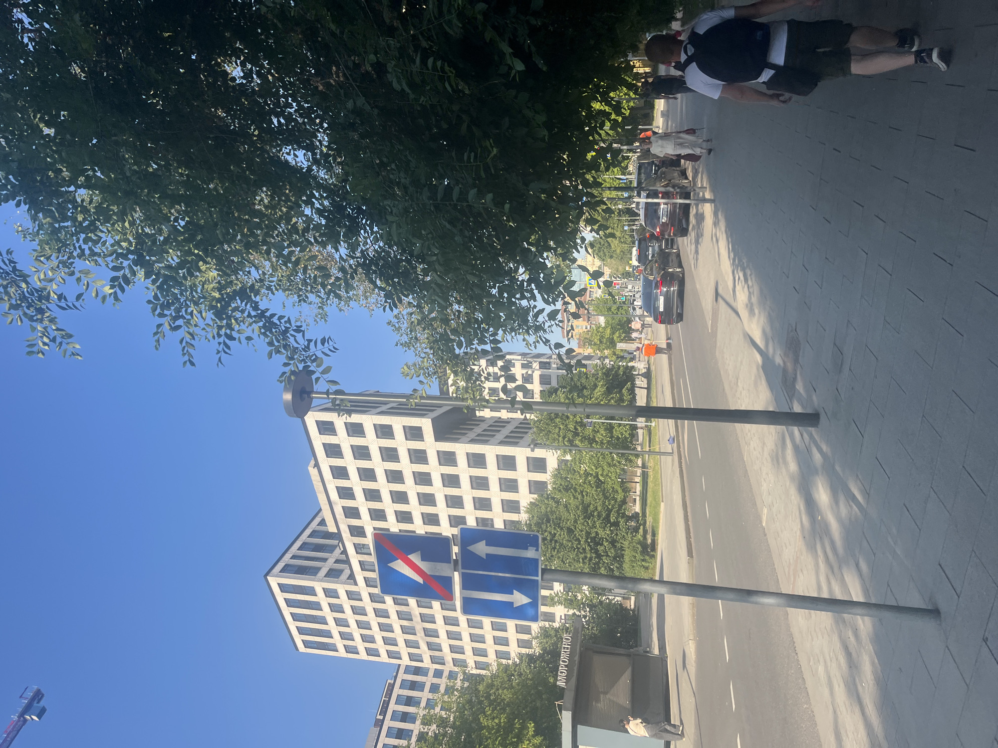 The image depicts a sunny day on a city street in Moscow, Russia. In the foreground, a man with a backpack walks away from the camera down a paved sidewalk. To the left, cars drive on a multi-lane road. Traffic signs are visible on the side of the road, indicating directions and lane restrictions. Behind the road, modern white buildings with dark windows stand tall, suggesting an urban business or residential area. Trees line the sidewalk and the median, providing shade. A kiosk with the word "МОРОЖЕНОЕ" (ice cream) written on it is visible to the left. The sky is clear and blue.