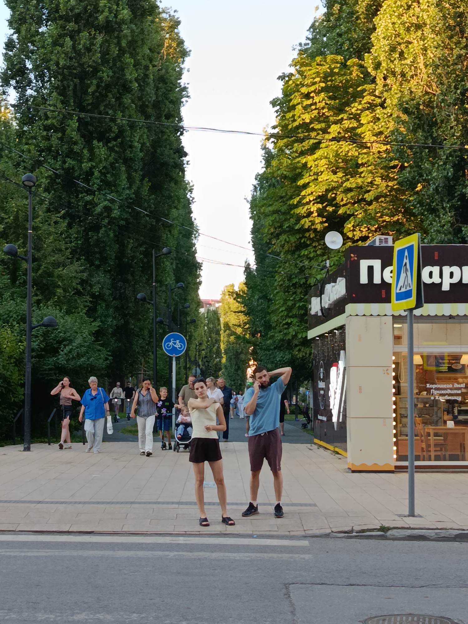 In Saratov, Russia, this image captures a street scene with pedestrians walking along a paved pathway lined with tall trees. A man in a blue t-shirt and brown shorts stands on the right, holding his phone to his ear and touching his head. A woman in a light-colored tank top and dark shorts stands in front of him, also touching her head. Behind them, other people are walking, some are on scooters, and a child is in a stroller. A kiosk with a yellow pedestrian crossing sign is visible on the right side of the pathway. The overall environment suggests a public space, possibly a park or a pedestrian street, bathed in the warm light of late afternoon or early evening.