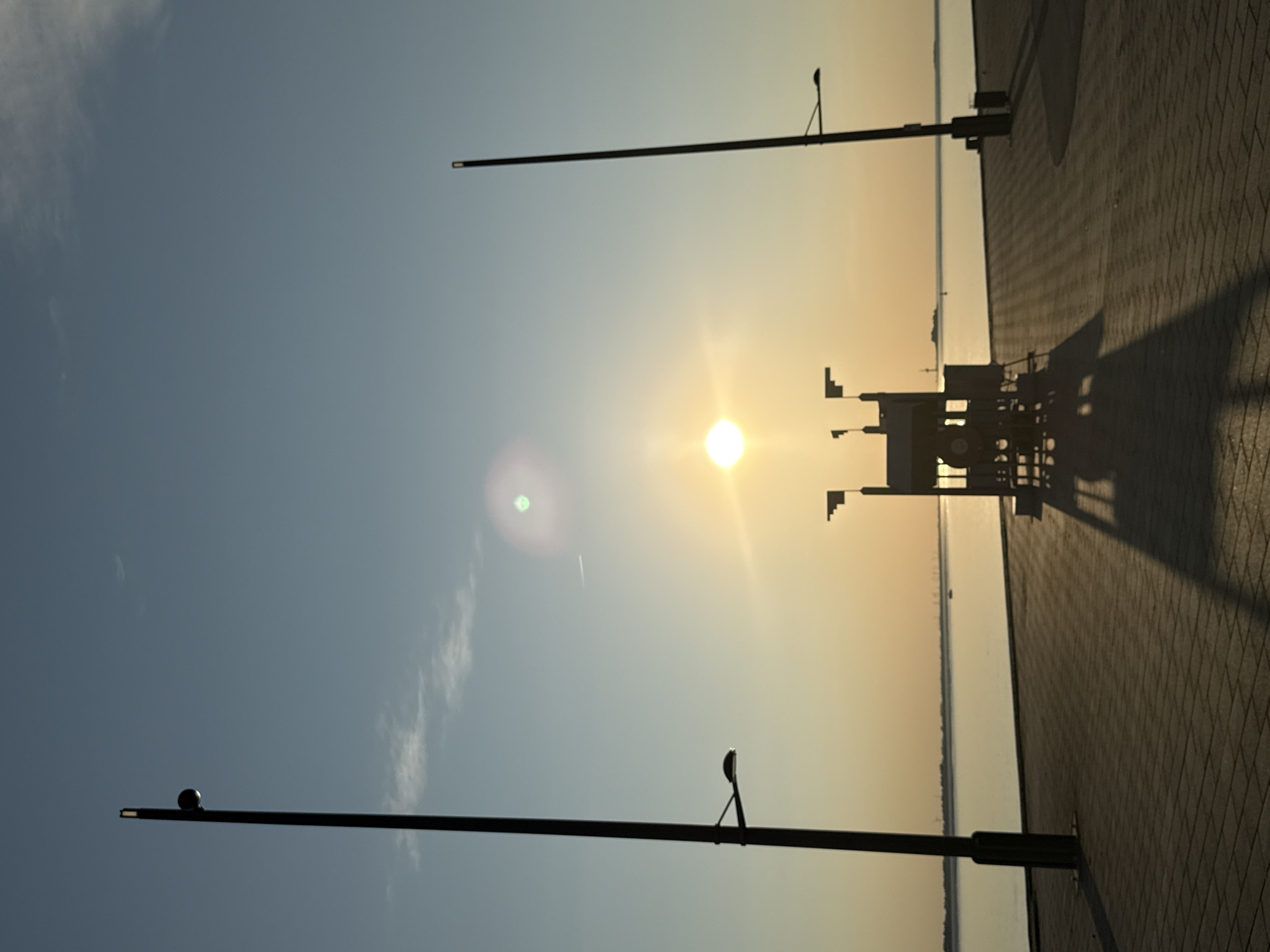 This is a vertical, eye-level shot of a waterfront promenade at sunset. The sky is a gradient of pale blue at the top, transitioning to a warm orange and yellow as it meets the horizon. The sun is visible as a bright, slightly hazy orb in the upper center of the frame, casting a lens flare.
In the foreground, the promenade is paved with rectangular stones laid in a patterned brick formation. Two tall, dark lamp posts stand on either side of the frame, casting long shadows across the promenade. The lamp post on the left is closer and taller, with two lights attached at different heights. The lamp post on the right is further away and thinner.
In the middle ground, a dark, abstract structure with flags or banners on top sits on the promenade, also casting a long shadow. It appears to be some sort of public amenity or art installation. Beyond the promenade, a calm body of water stretches to the horizon, reflecting the warm colors of the sky. A distant shoreline with trees and structures is faintly visible on the horizon, along with a few small boats or buoys on the water. The overall mood is serene and tranquil, with the setting sun creating a warm and inviting atmosphere.