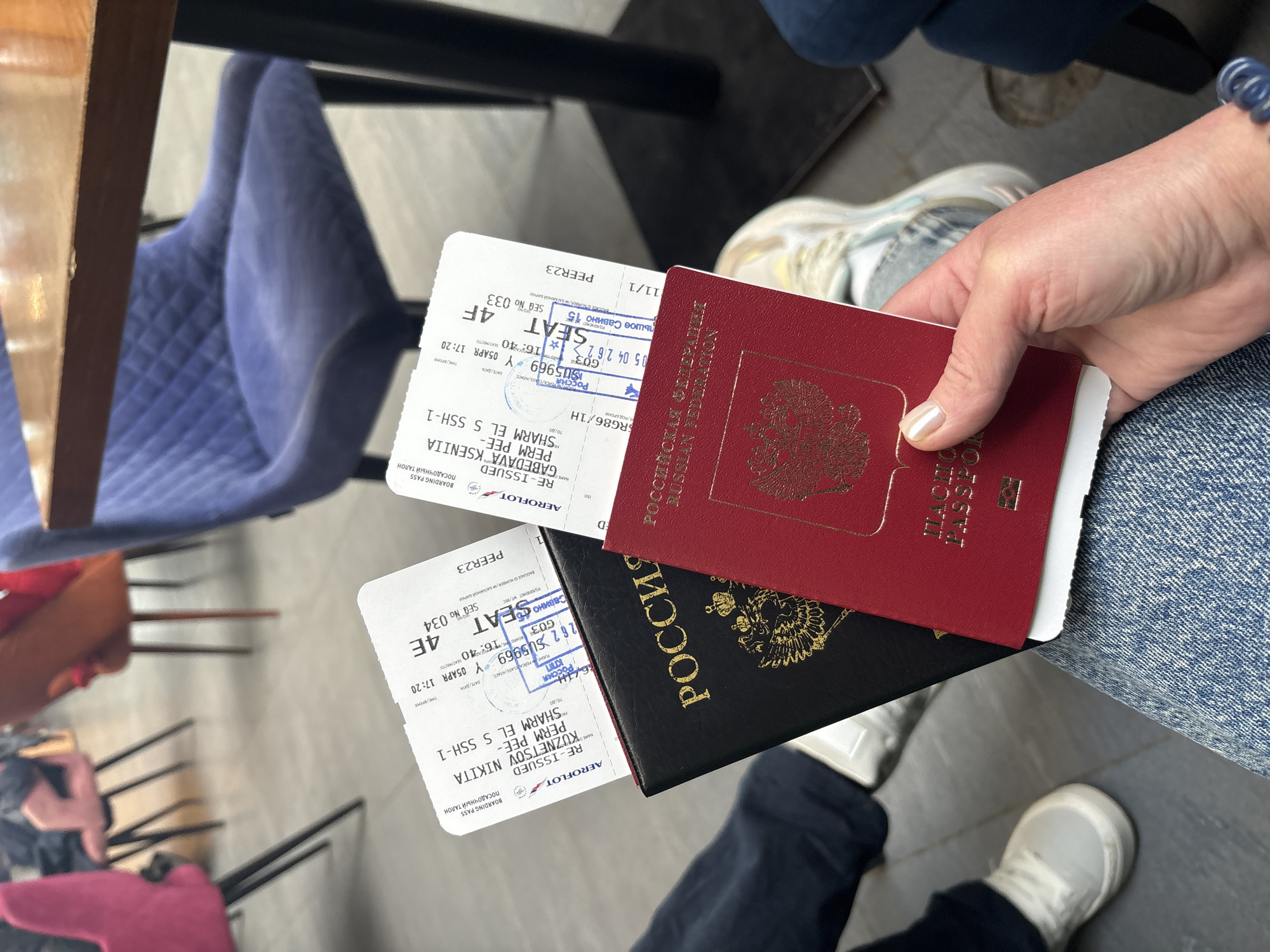 A person is holding two passports and two boarding passes. The passports are Russian, one is red and one is black. The boarding passes are both for Aeroflot flights, with seat 4E. One pass is for a flight from Perm to Sharm el-Sheikh and the other is for a flight from Perm to Sharm el-Sheikh. The boarding passes also mention Bolshoye Savino as the departure location. The date and time on both boarding passes are April 5, 17:20. The person is wearing jeans and sneakers. A blue velvet chair can be seen in the background.