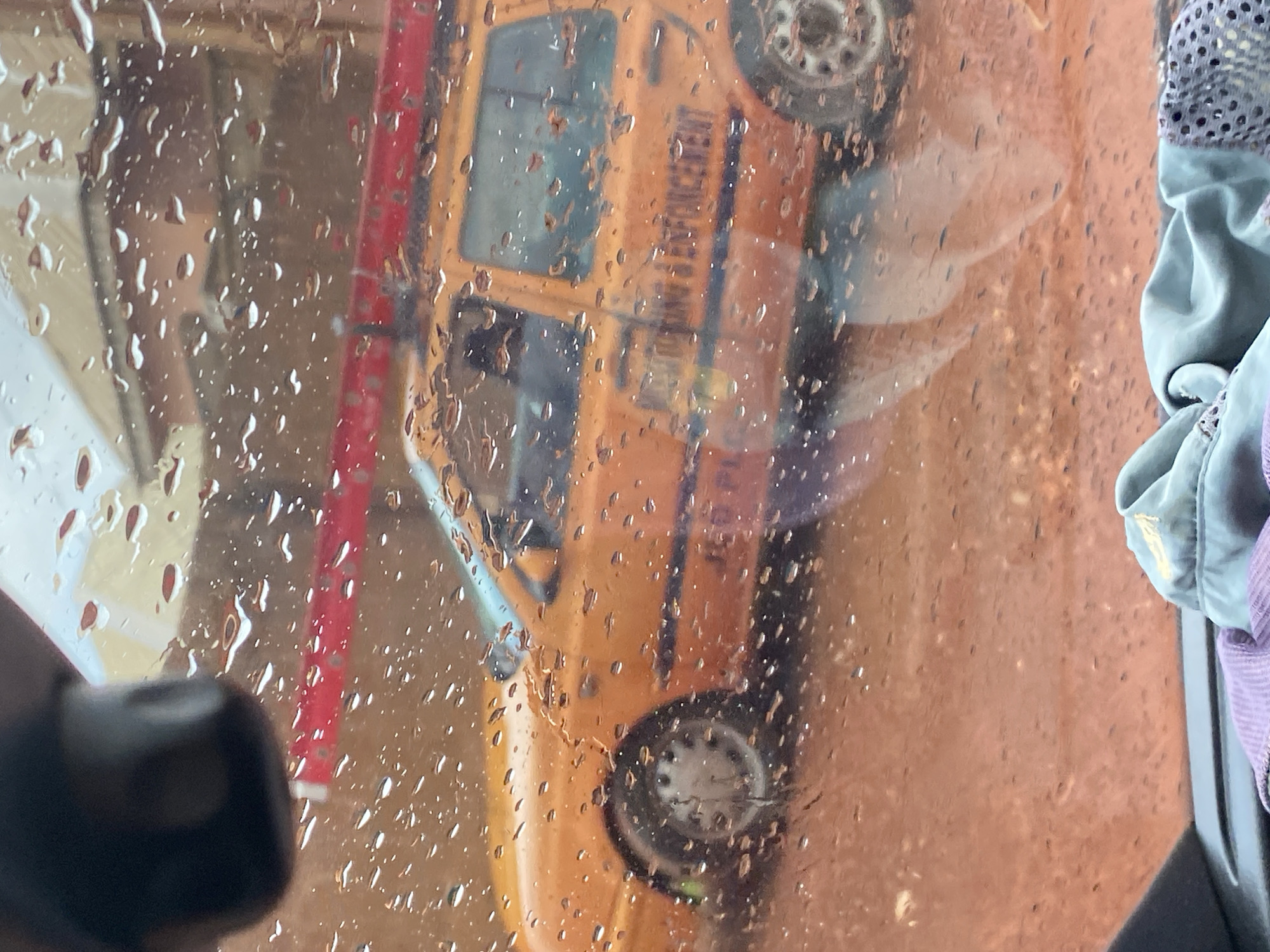 The image captures a close-up view of a window, likely from inside a vehicle, covered in numerous water droplets, indicating rainy weather. Through the rain-streaked window, an orange vehicle, possibly a school bus or a utility vehicle, is visible. The vehicle appears to be in a street or parking lot. Reflections on the wet glass distort the view, creating a slightly abstract and impressionistic scene.
A portion of a red pole or barrier is seen to the left of the orange vehicle. The text "LAW ENFORCEMENT" is partially legible on the side of the orange vehicle, suggesting it is used by some form of law enforcement. A grey or blue fabric, possibly part of a bag or clothing, is visible in the upper right corner of the image, draped over the edge of the window frame. The lighting suggests it could be daytime, but the overcast sky due to the rain makes it difficult to determine the exact time. The scene evokes a sense of being indoors, observing the outside world through a wet and distorted lens, perhaps during a commute or while waiting. The overall mood is somewhat muted, consistent with rainy conditions. The location context of Jos, Nigeria, is provided, but there are no specific visual cues in the image itself to confirm this location.