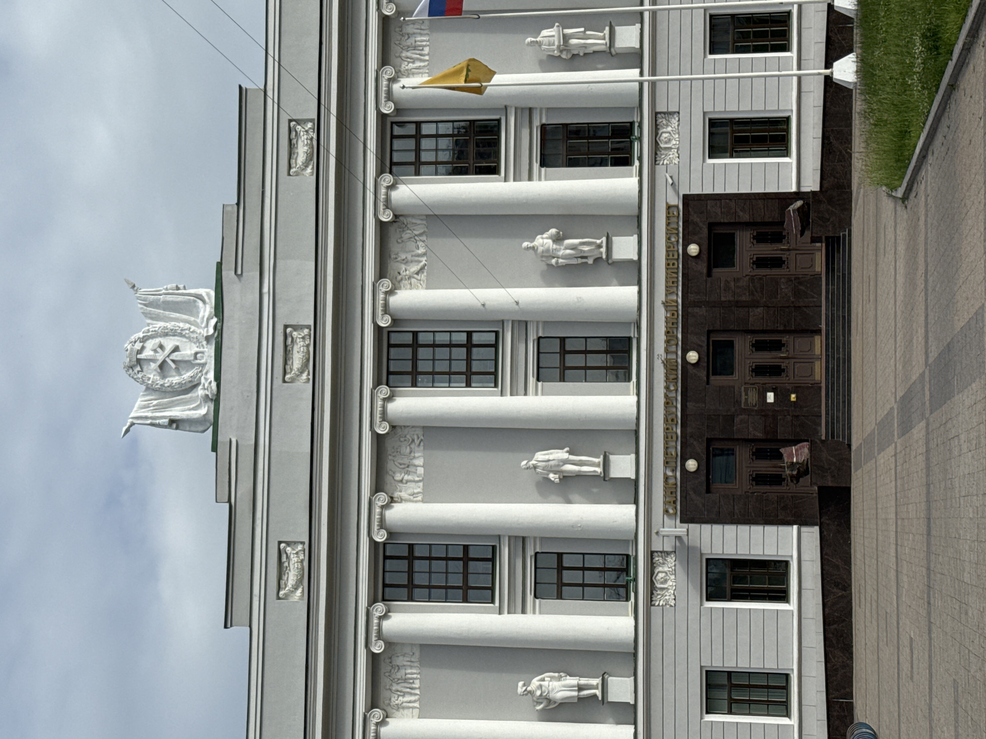 The image shows the facade of the Saint Petersburg Mining University, located in Saint Petersburg, Russia. The building is a large, classical-style structure with a grey facade, white columns, and a pediment adorned with a sculpture. Statues of historical figures are placed along the facade. In front of the building is a paved courtyard, and to the right, there is a grassy area. Two flags, one yellow and one tricolor, are visible on flagpoles in front of the building. The sky is overcast.