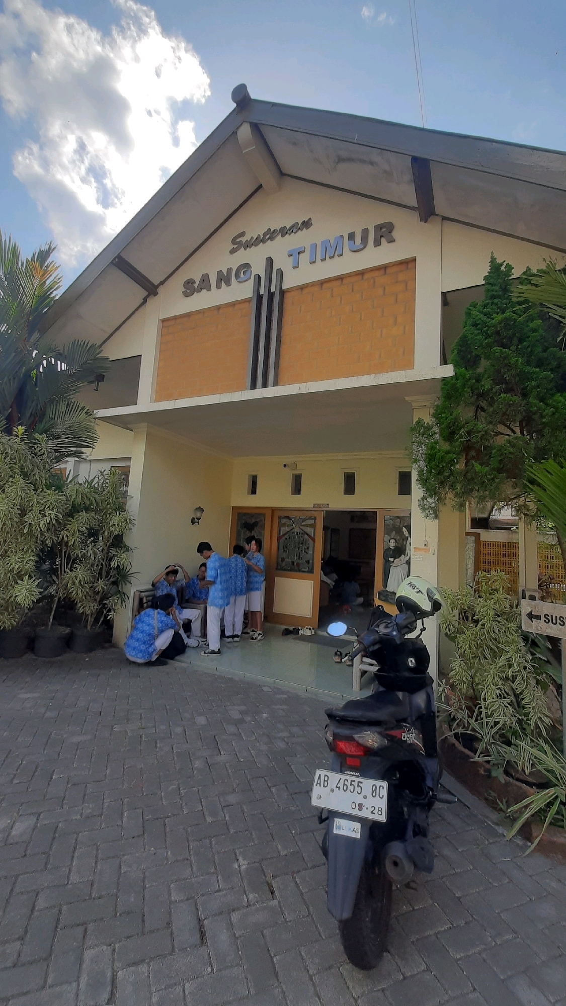 A medium-shot exterior photograph captures the front entrance of a building labeled "SUSTERAN SANG TIMUR" in Nanggulan, Indonesia. The building has an angled roof with dark gray trim and a light-colored facade. The upper portion of the entrance features orange brickwork with the building's name in silver, raised lettering. Below the name, three dark vertical accents are visible. The entrance itself is recessed and has double wooden doors with decorative glass panels.
Several young people, appearing to be students wearing blue and white uniforms, are gathered on the porch and in front of the entrance. Some are sitting on the ground, while others are standing and interacting. A black motorcycle with a white helmet on the handlebars is parked in the foreground on the brick-paved walkway leading to the building. To the right of the entrance, a sign with an arrow points to "SUS" (likely the beginning of a word like "SUSTERAN" or a related term). Lush green plants in dark pots are situated around the entrance and to the left of the building. The sky above is partly cloudy with a bright sun.