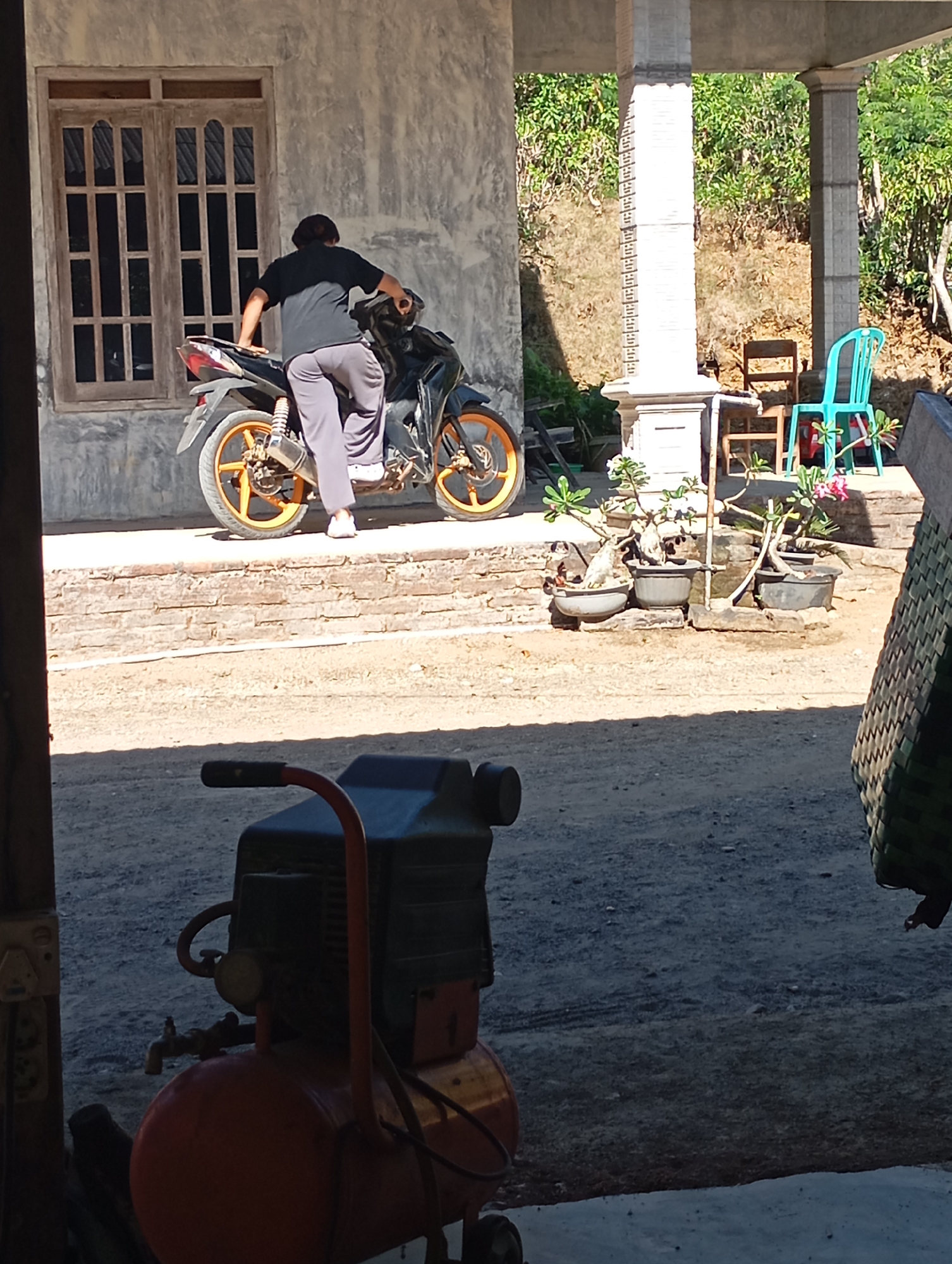 In Ketepung, Indonesia, a person in a black t-shirt and gray pants sits on a black motorcycle with orange rims. The motorcycle is parked in front of a building with a textured gray wall and a wooden-framed window with multiple panes. To the right, a pillar supports an overhang, and beyond that, a bright turquoise plastic chair is visible, along with wooden chairs and potted plants, including a flowering plant with pink blossoms. The ground is a mix of dirt and gravel. In the foreground, partially obscuring the lower portion of the scene, is a red and black air compressor with various hoses and attachments. The background features dense green foliage on a slope.