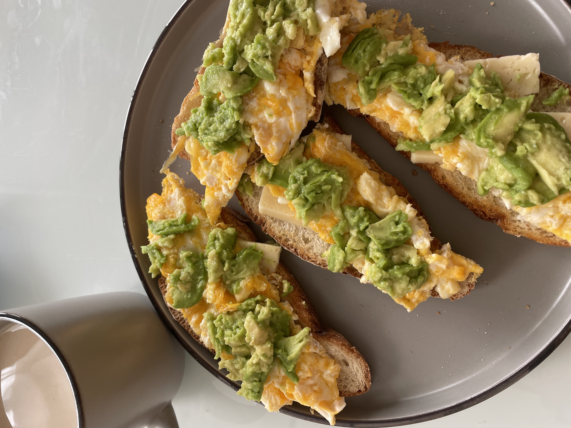 An overhead medium shot captures a rustic breakfast spread on a gray plate. Four slices of toasted artisanal bread form the base for creamy scrambled eggs, melted cheese, and a generous topping of diced avocado. A mug filled with a light-colored beverage, likely coffee or tea, sits to the left, its rim lined with a darker shade. The setting appears to be a home kitchen or dining area, with a smooth, light-colored surface extending beyond the plate.