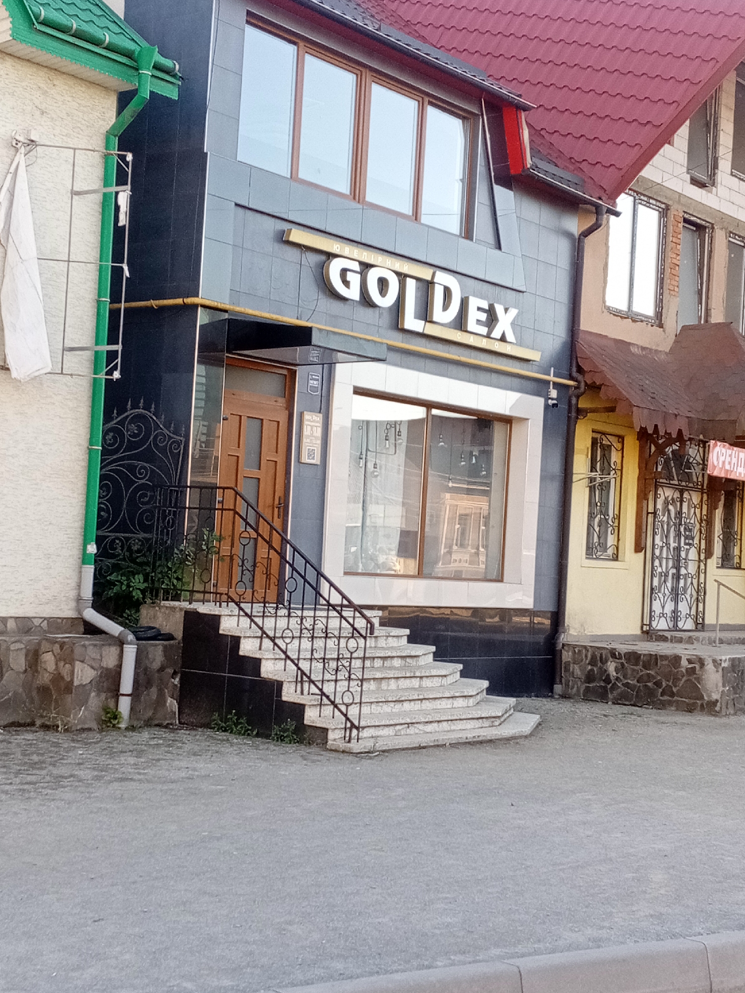 This is a medium-shot, eye-level, outdoor, daylight photograph of the facade of a commercial building in Tiachiv, Ukraine. The building is a multi-story structure with a prominent storefront. The main facade visible is dark gray with a large sign featuring the text "GOLDEX" in white, three-dimensional letters. Below "GOLDEX" is smaller text that appears to read "САЛОН". The storefront itself has large windows. To the left of the main facade, a portion of an adjacent building is visible, painted white with a green roofline and a green downspout. To the right of the "GOLDEX" storefront, another section of the building is visible, painted yellow with a dark, weathered awning over a doorway with a decorative metal gate. A red sign with white text, possibly indicating "SALE" or "RENT" in Cyrillic, is attached to the yellow section. The buildings are situated on a paved area, possibly a sidewalk or pedestrian walkway, with a cobblestone or stone foundation visible at the base of the structures. A set of concrete stairs with a black metal railing leads up to the "GOLDEX" entrance. The overall scene depicts a commercial street or area in a Ukrainian city. There are no people visible in the image.
