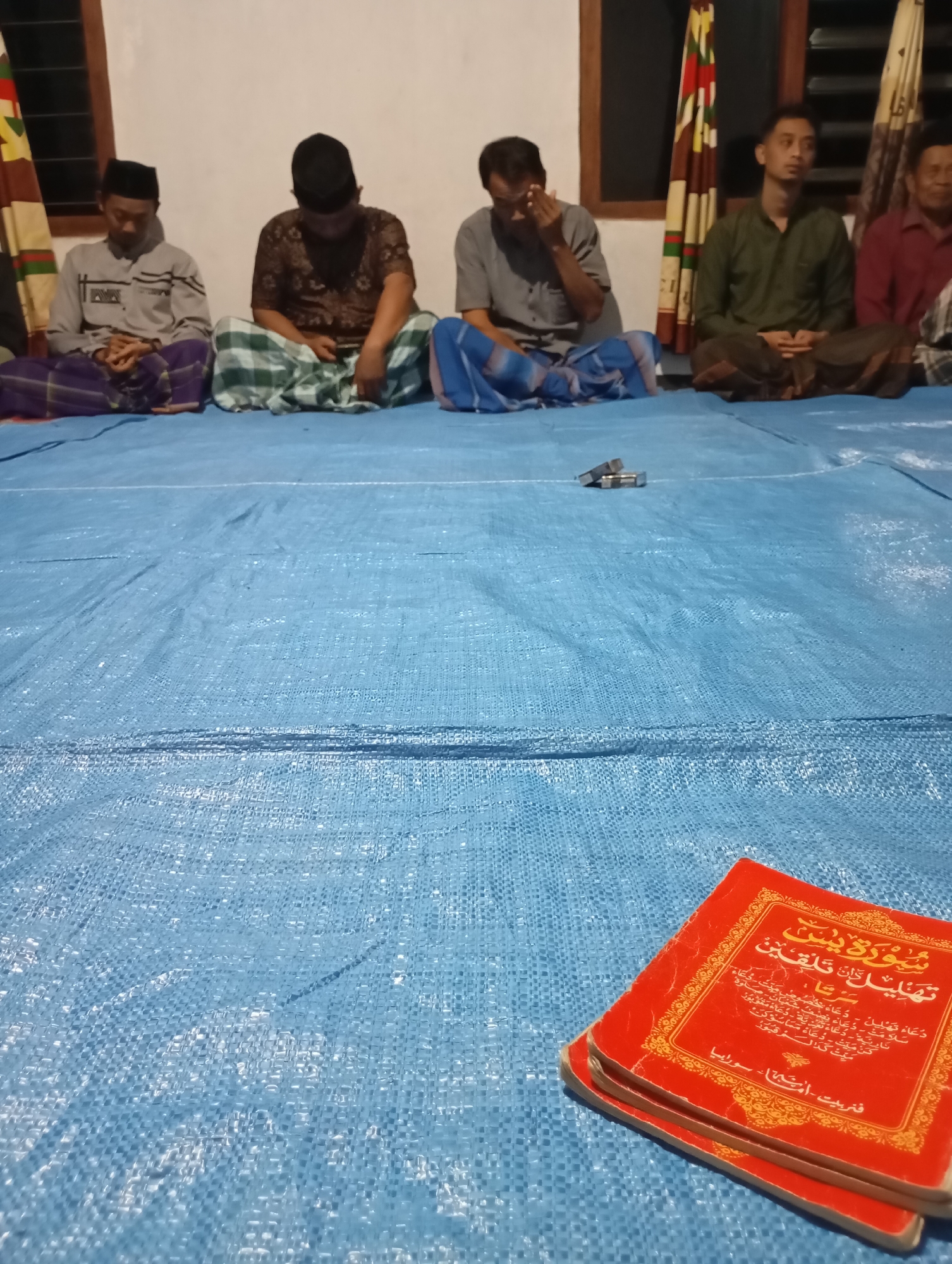 In Ketepung, Indonesia, five men are seated cross-legged on a blue tarpaulin on the floor. The men are dressed in traditional Islamic attire, including peci (caps) and sarongs. Some appear to be engaged in prayer or religious reflection. In the foreground, a stack of red books with Arabic script is visible. The setting appears to be an indoor space, possibly a home or a place of worship, with a window and curtains visible in the background. The lighting suggests it is evening or nighttime.